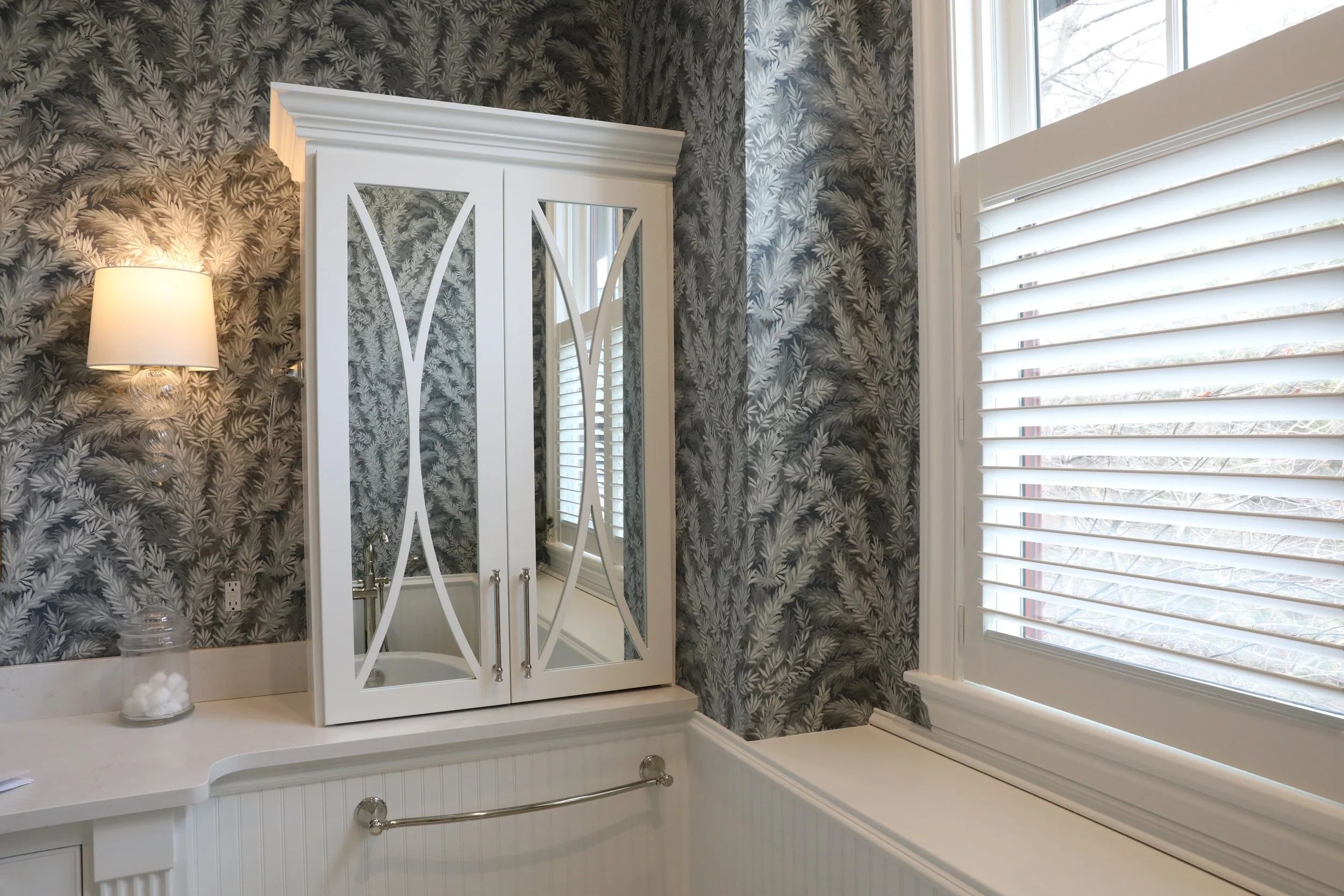 A bathroom corner featuring a bathtub, a mirror cabinet, a wall-mounted lamp, and a window with white blinds. The walls are decorated with dark, leafy patterned wallpaper.