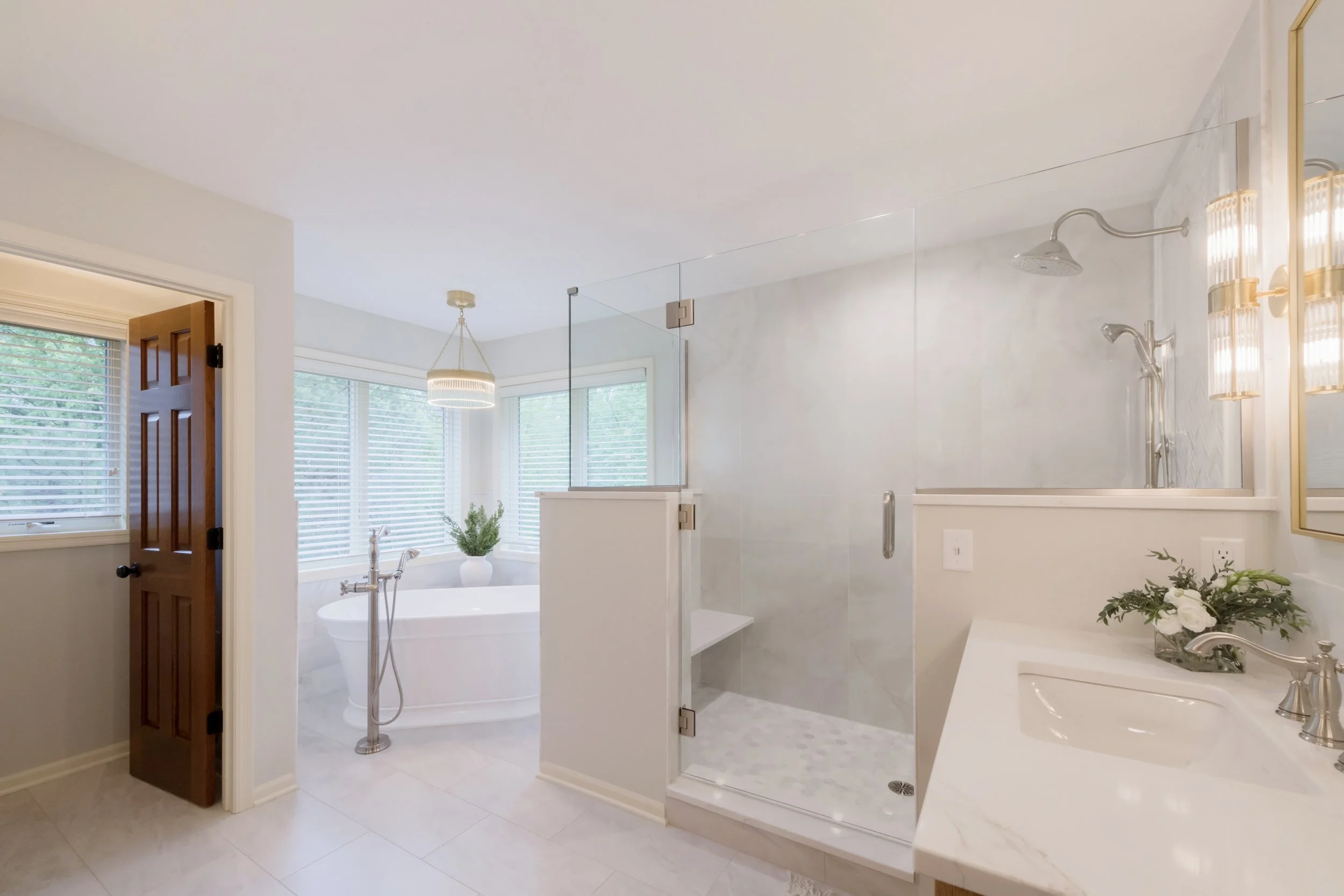 A bright and white bathroom featuring a walk-in shower with glass enclosure, a bathtub near windows with white blinds, a ceiling light fixture, a wooden door, a sink with a mirror, and a small table with a flower arrangement.