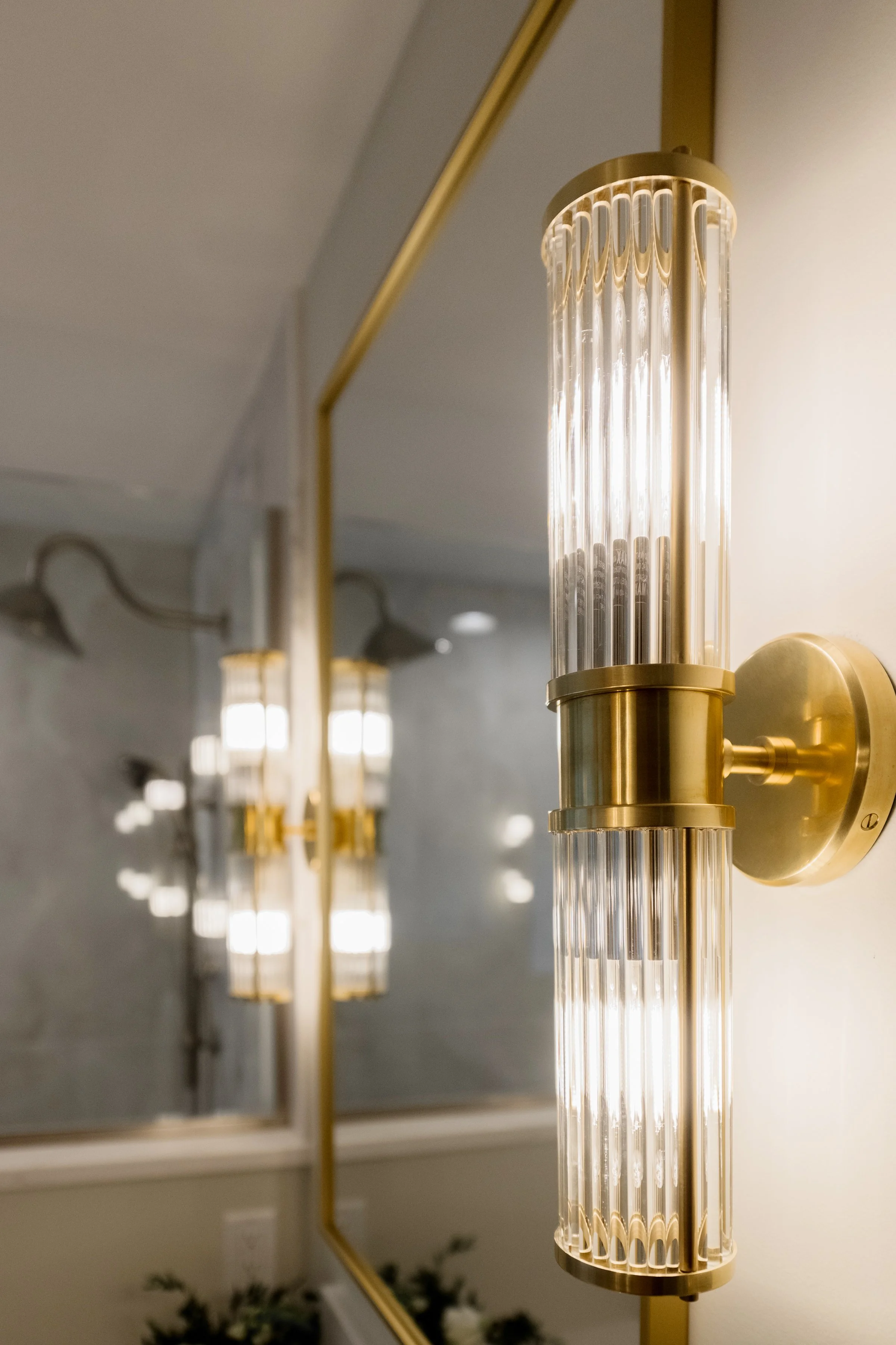 A modern gold wall-mounted lamp with a cylindrical glass shade, positioned in front of a mirror.
