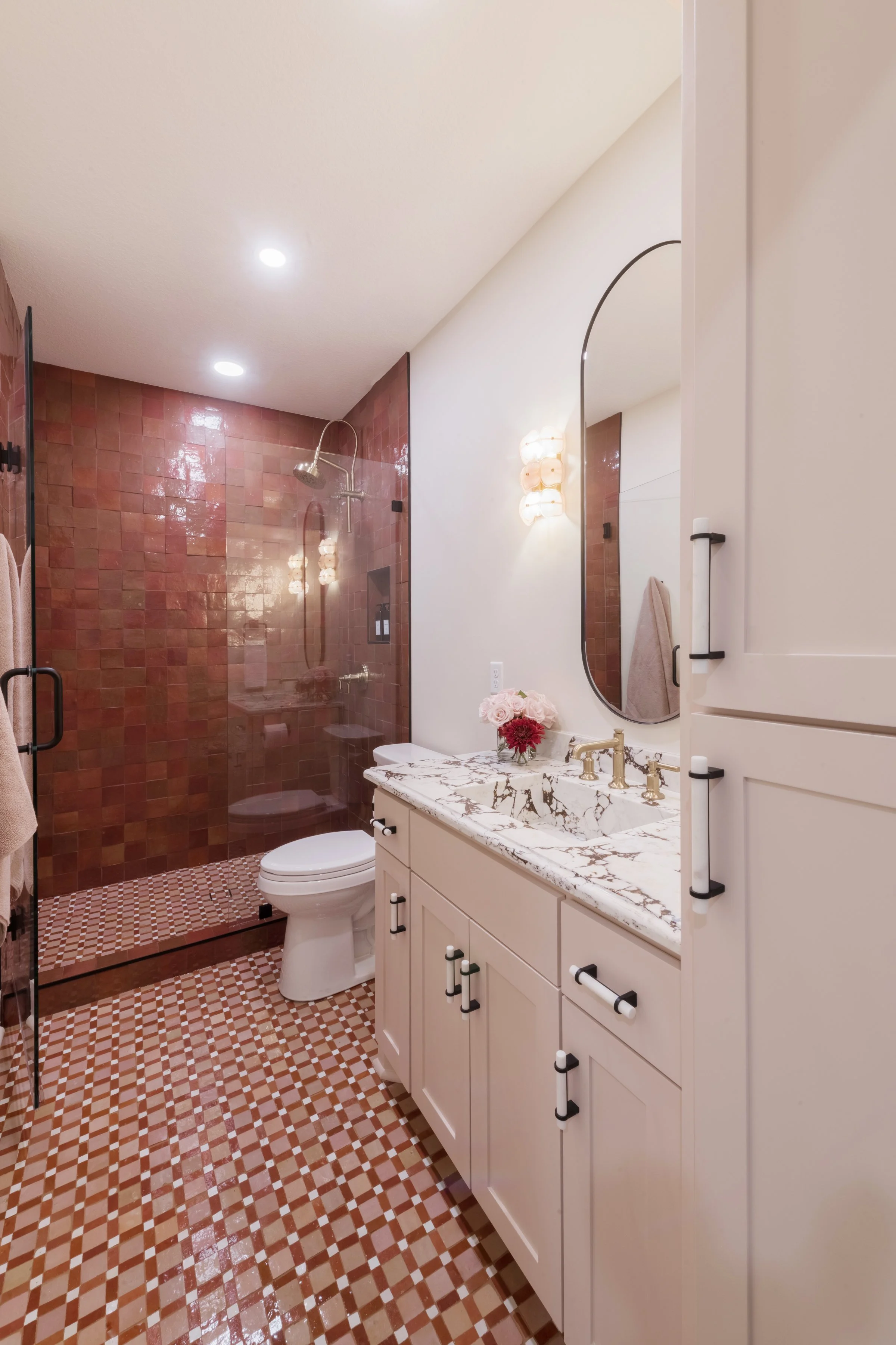 A bathroom with a shower area featuring pinkish-red tiles, a glass shower door, a white toilet, and a vanity with a marble countertop, a mirror, and decorative flowers.