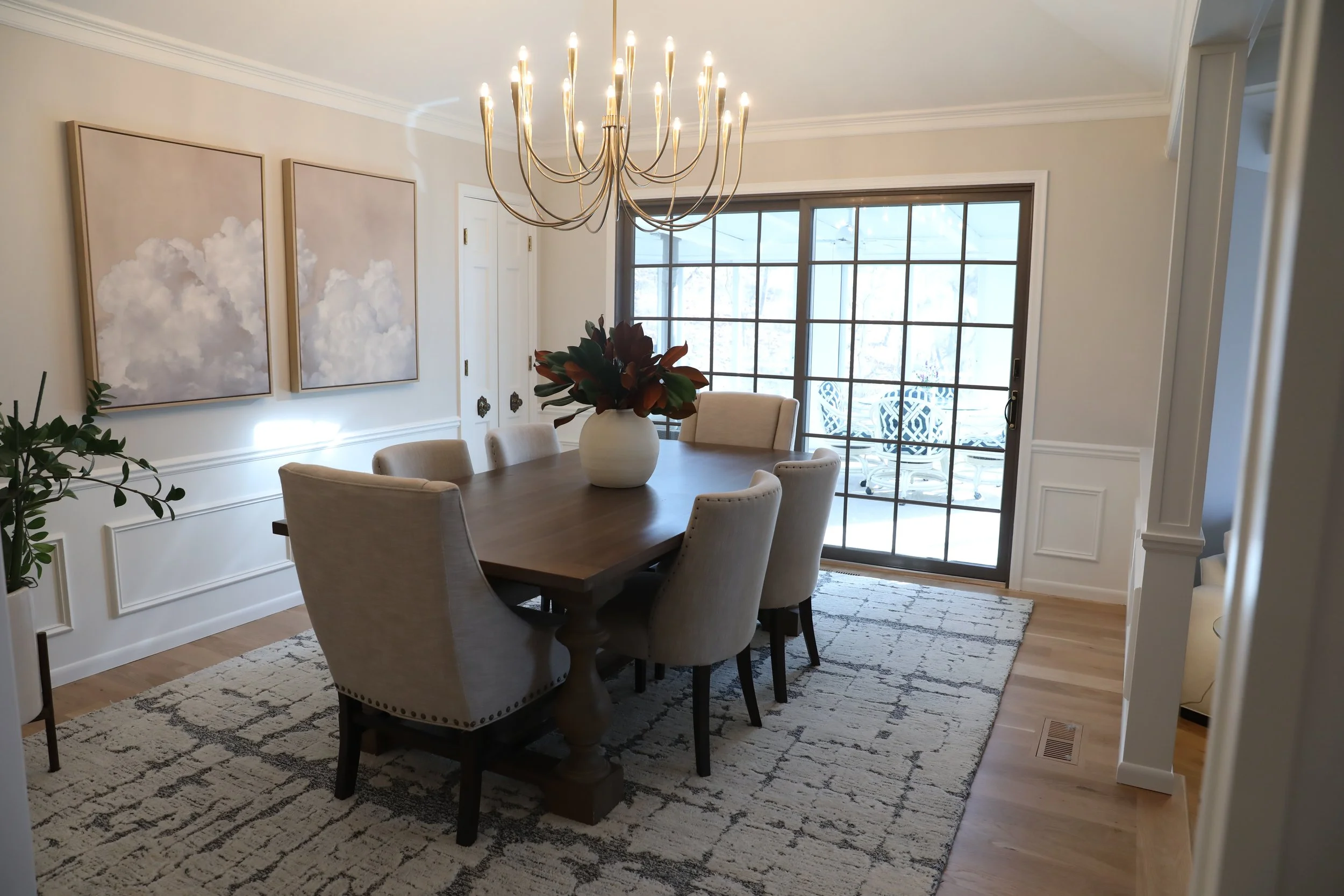 A dining room with a wooden table surrounded by six cream-colored upholstered chairs, a white vase with dark green and red plants, a patterned rug, two large paintings of clouds on the wall, a chandelier hanging above the table, a sliding glass door 