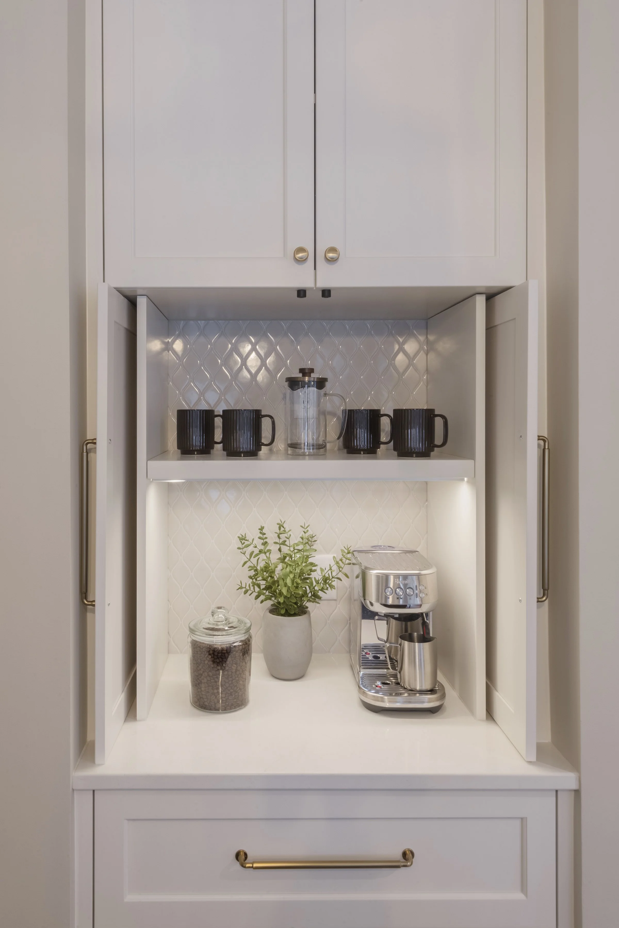 A white kitchen cabinet with an open middle section showing four black mugs, a glass coffee pot, and a small plant in a white pot. Below, a jar of coffee beans and an espresso machine are visible.