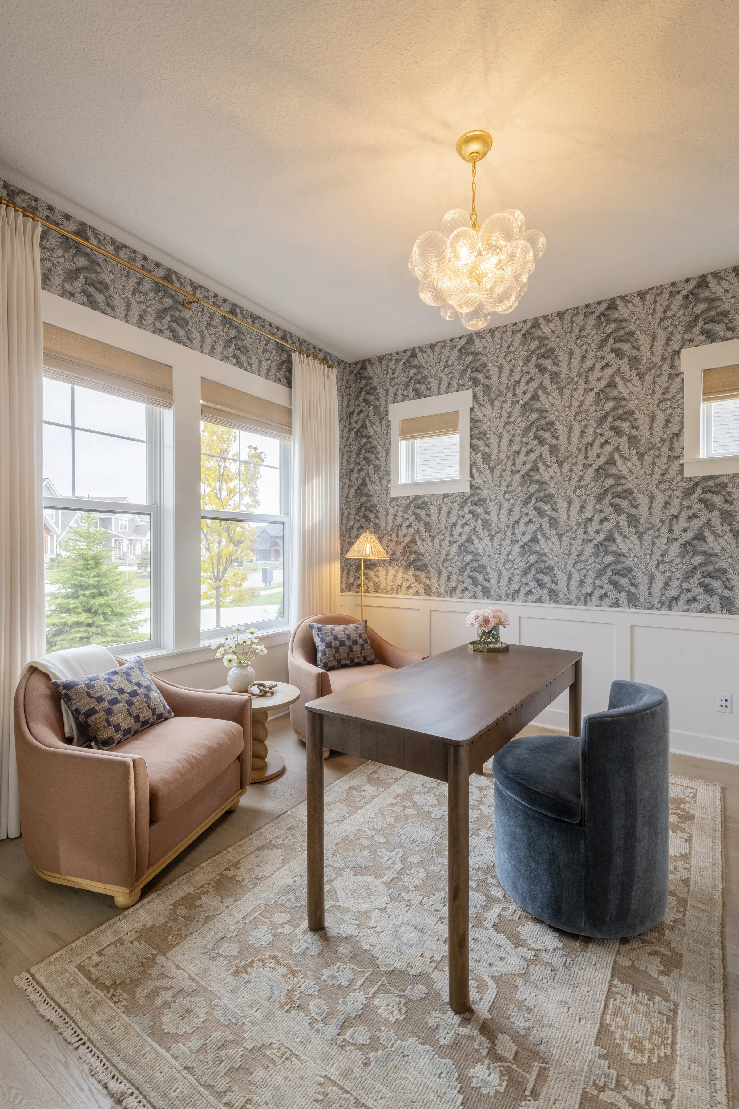 Living room with pink armchairs, a wooden table, a blue velvet chair, a patterned area rug, large windows with white curtains, a small table with a vase of flowers, a floor lamp, wallpaper with a leafy pattern, a chandelier, and a view of a suburban neighborhood outside.