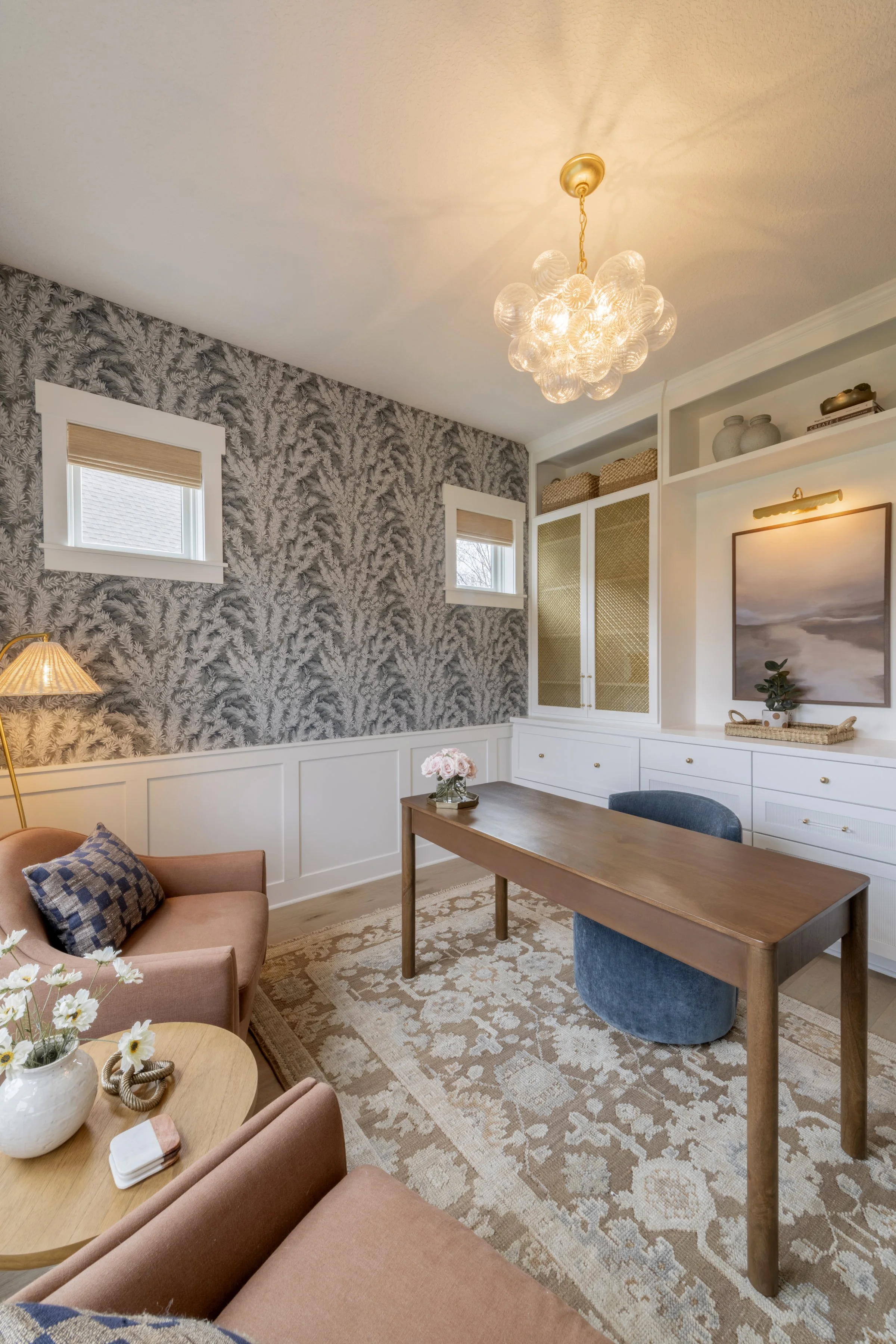 A cozy living room corner with a peach-colored armchair, a wooden side table with a white vase of flowers, a patterned beige and white rug, a wooden table, and a blue upholstered ottoman.