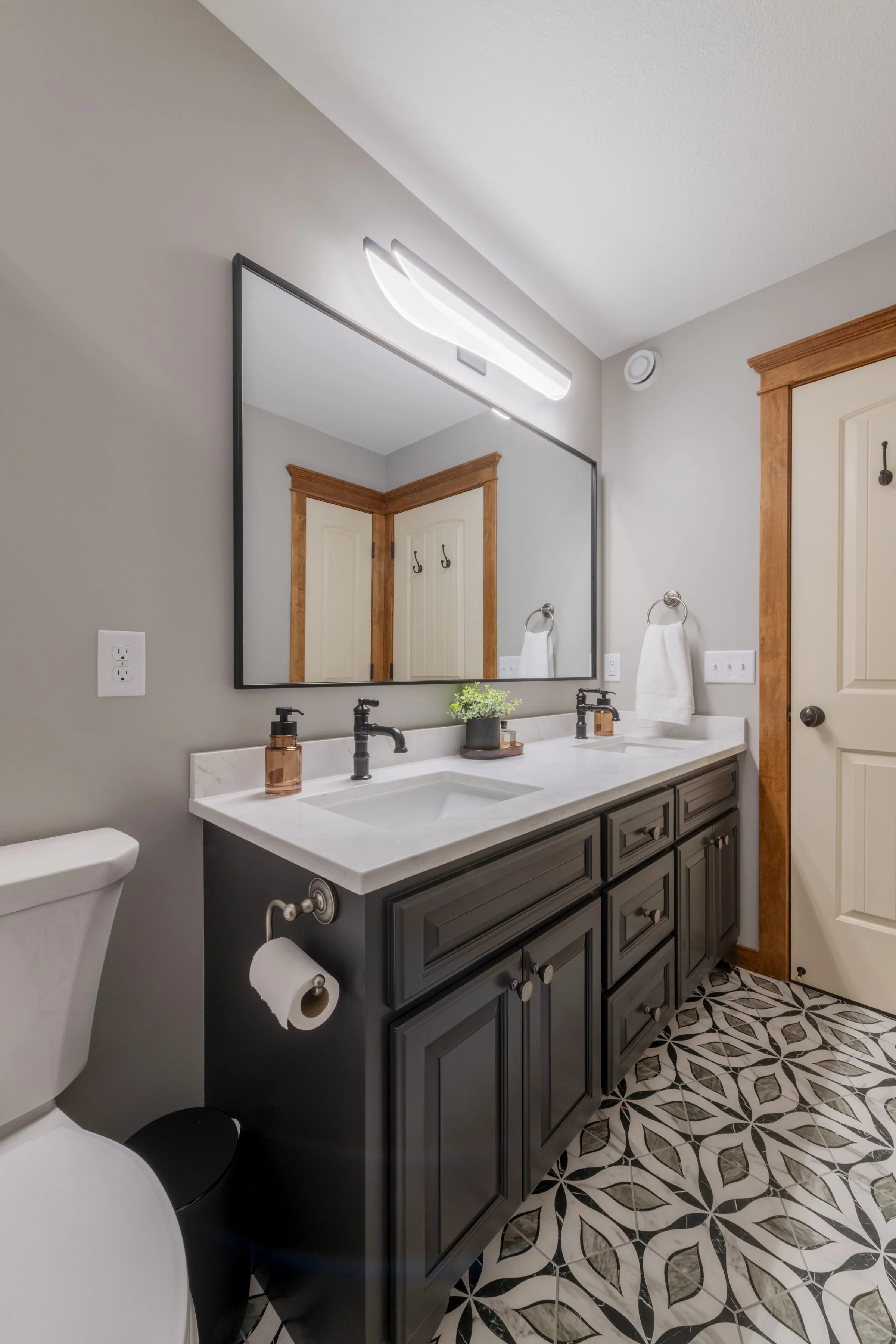 Bathroom with double sink vanity, large mirror, black fixtures, gray cabinets, decorative floor tiles, and wooden door frames.
