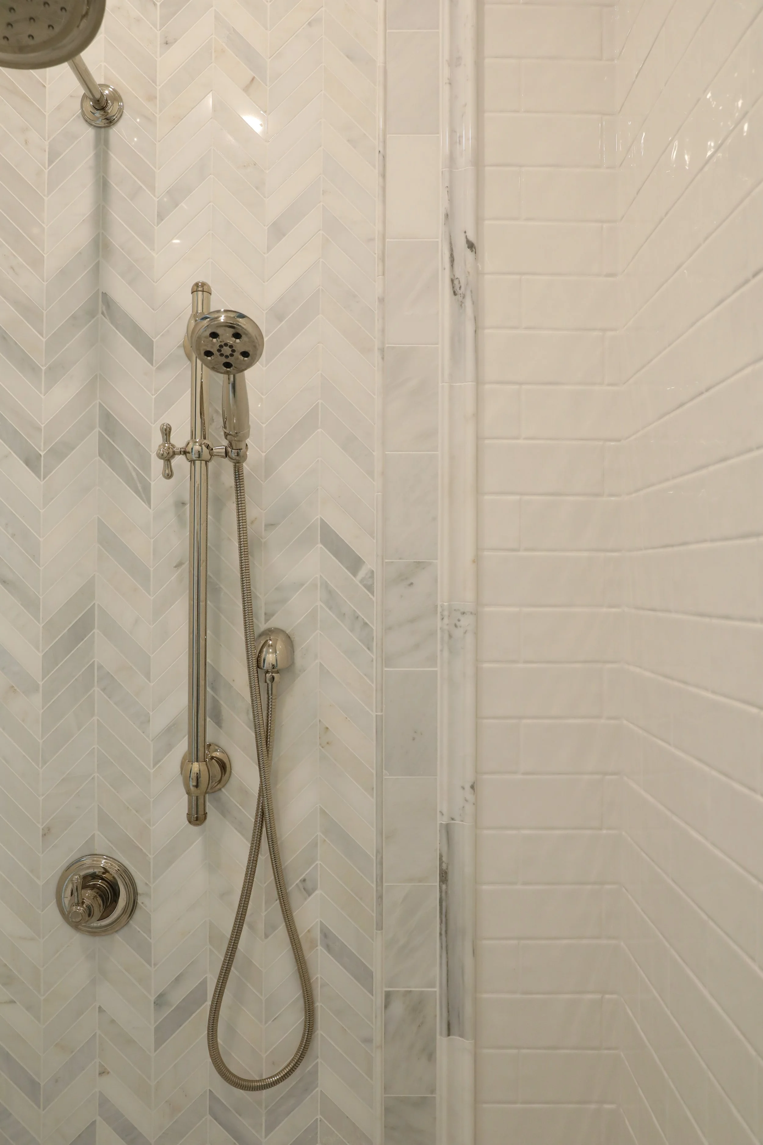 Close-up view of a shower with marble and herringbone pattern tiled walls, a handheld showerhead, and a vertical white pipe.