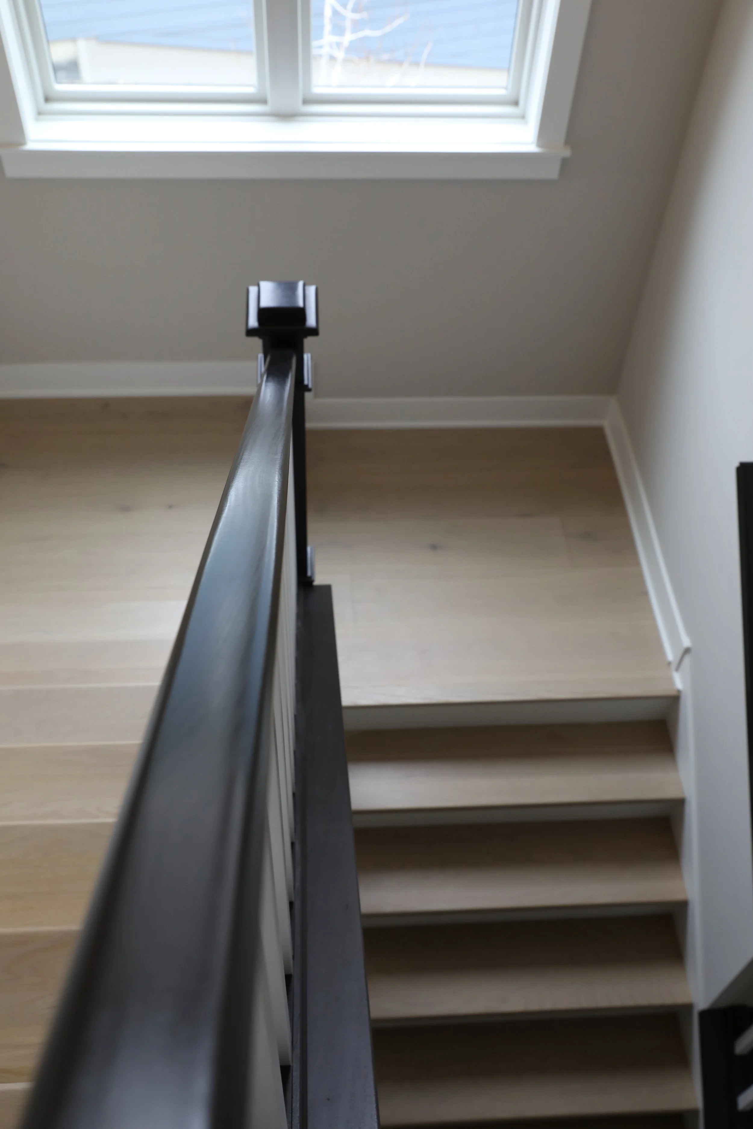 View from the top of a staircase looking down, with a black handrail and beige stairs, and a window at the top showing an exterior wall and some snow outside.