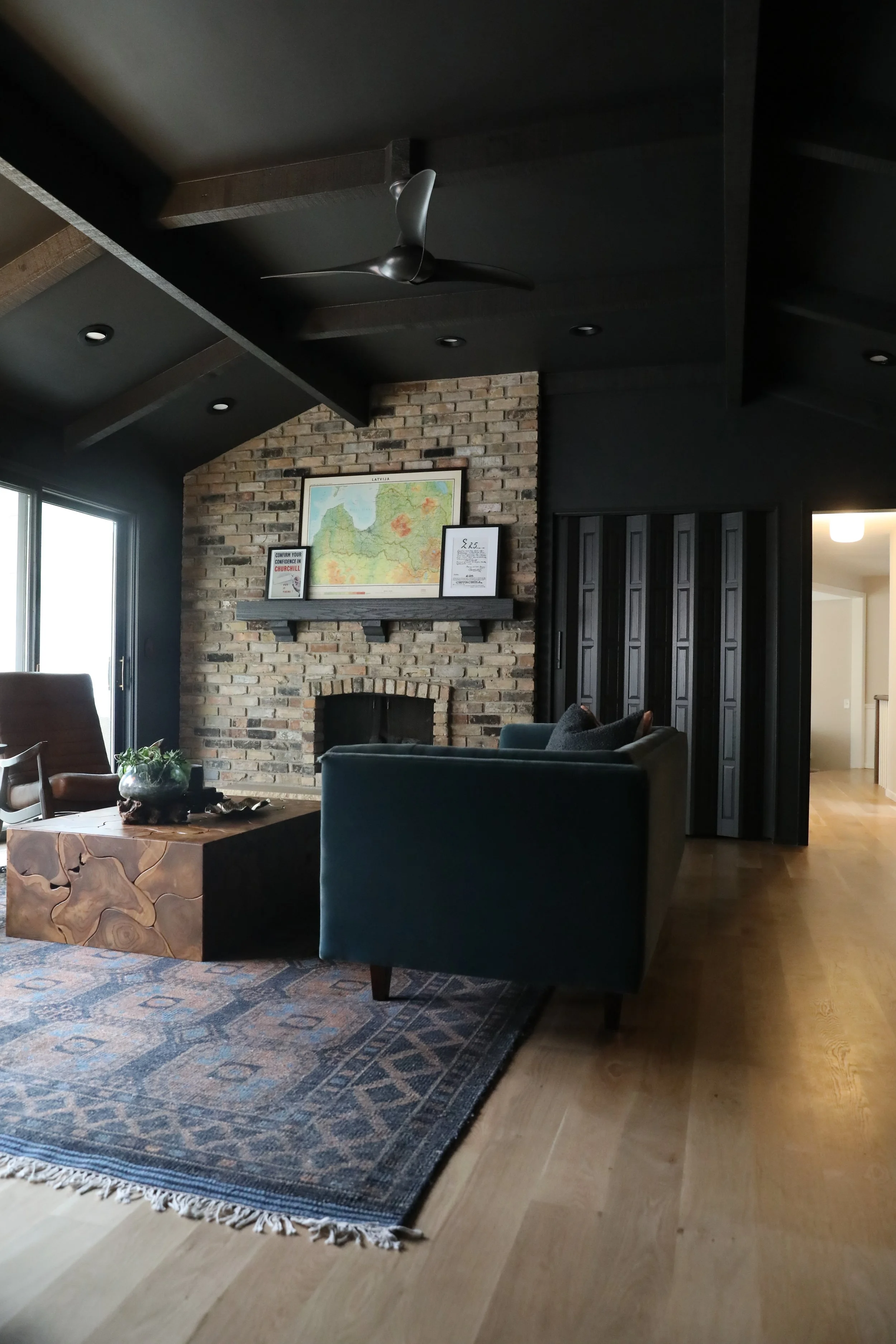 Living room with brick fireplace, mounted map, dark ceiling with recessed lights, wooden floor, dark furniture including a teal armchair and a wooden coffee table, and a sliding glass door.