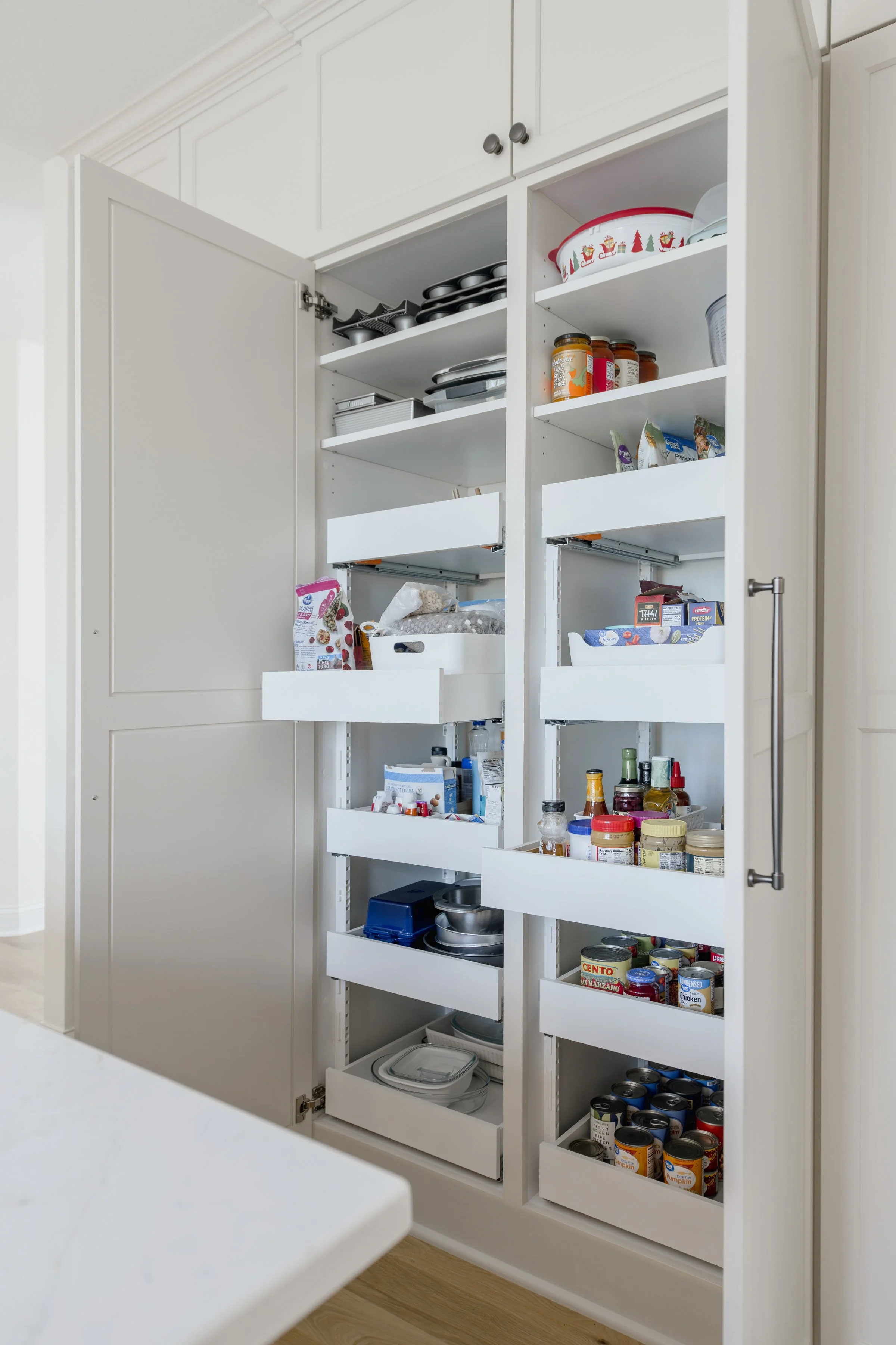 Open kitchen cabinet with multiple shelves containing canned goods, jars, boxes, and kitchenware.