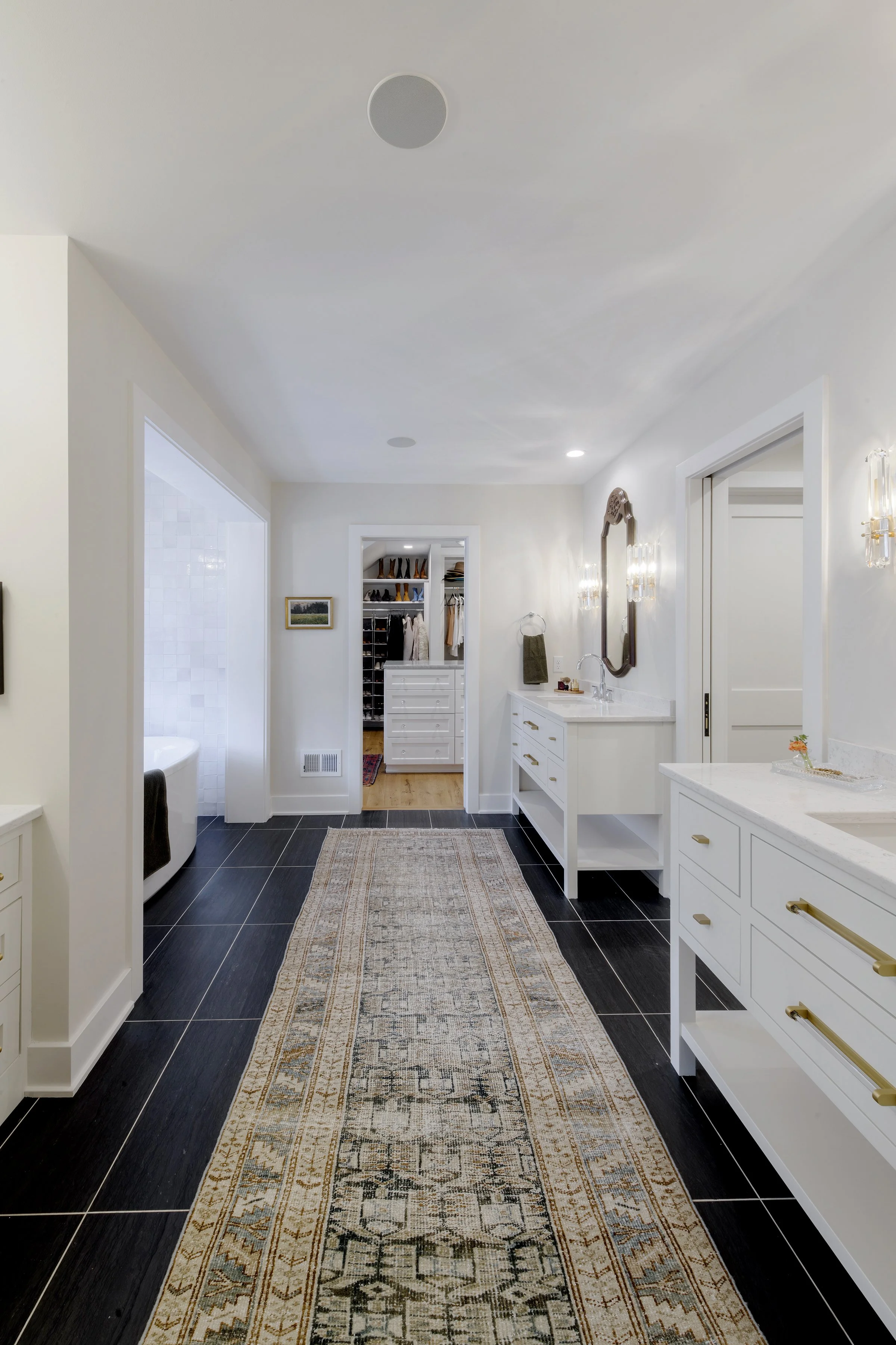 A spacious bathroom with white walls, black tile flooring, a white vanity with gold handles, a large mirror, and a long decorative rug. A walk-in closet with shelves and clothing is visible in the background.