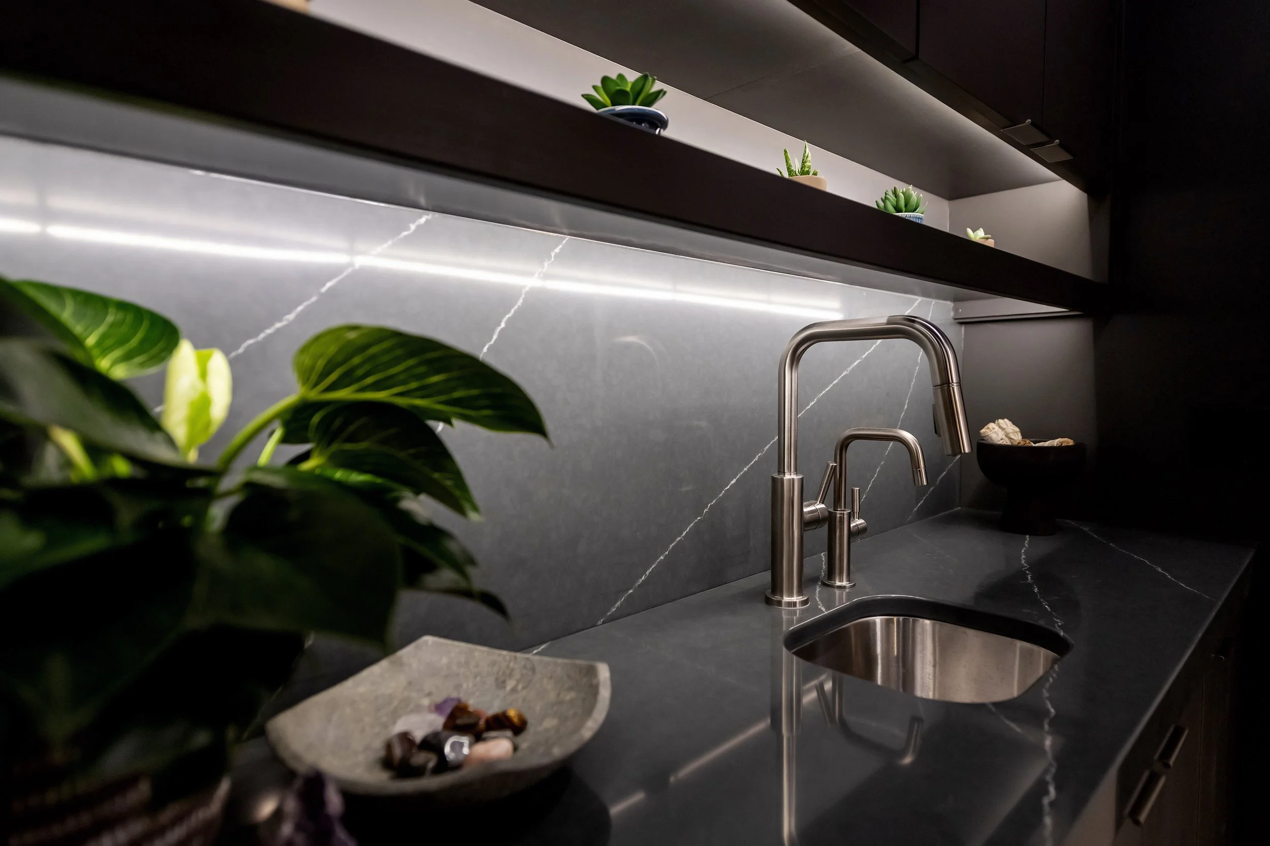 A modern kitchen sink with a city gray marble backsplash, stainless steel faucet, and green plants on open shelves above.