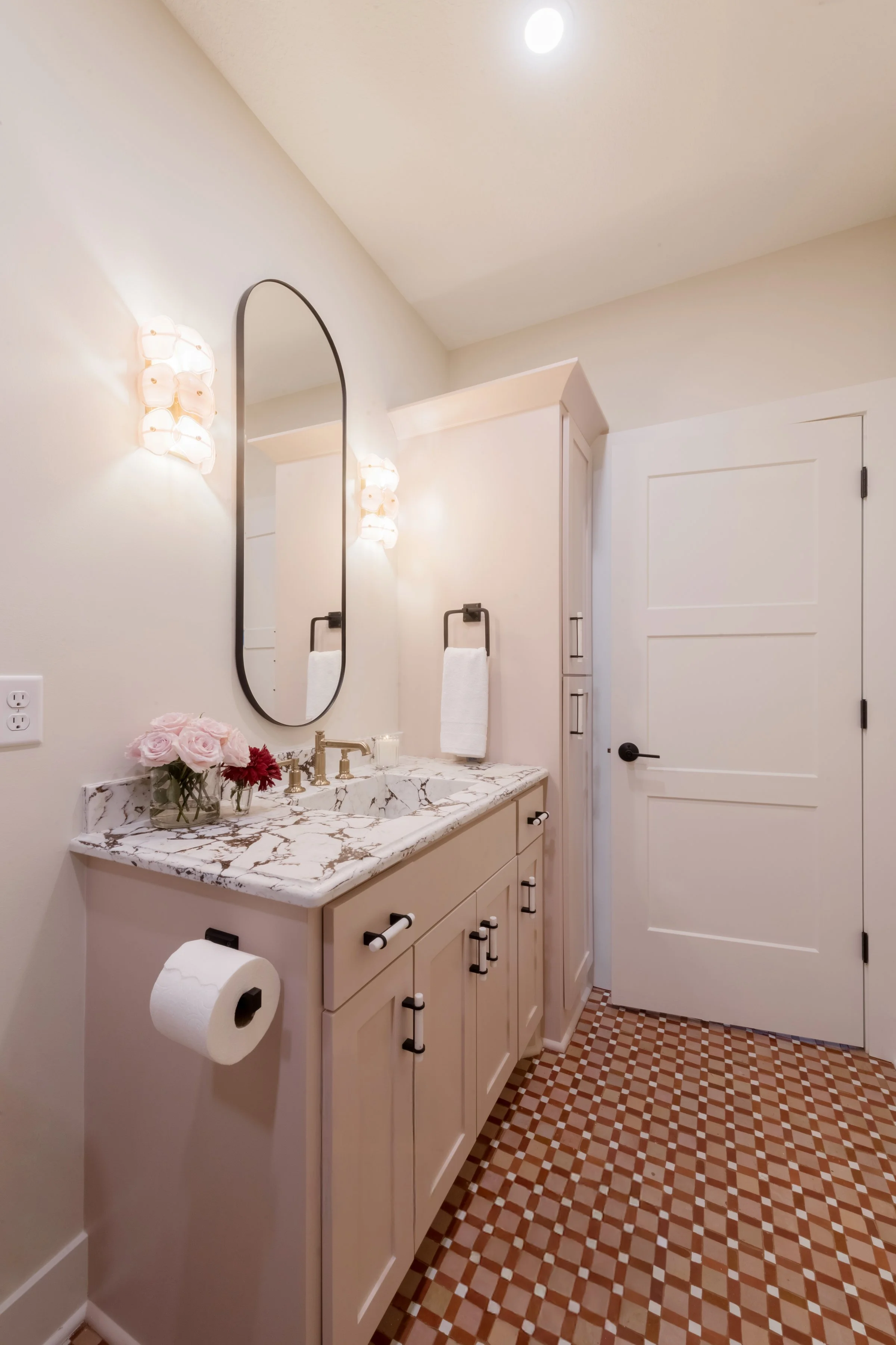 A bathroom with a white vanity with a marble countertop, a large oval mirror, pink flowers in a vase, wall sconces, a towel, a door, a toilet paper roll, and a patterned tiled floor.