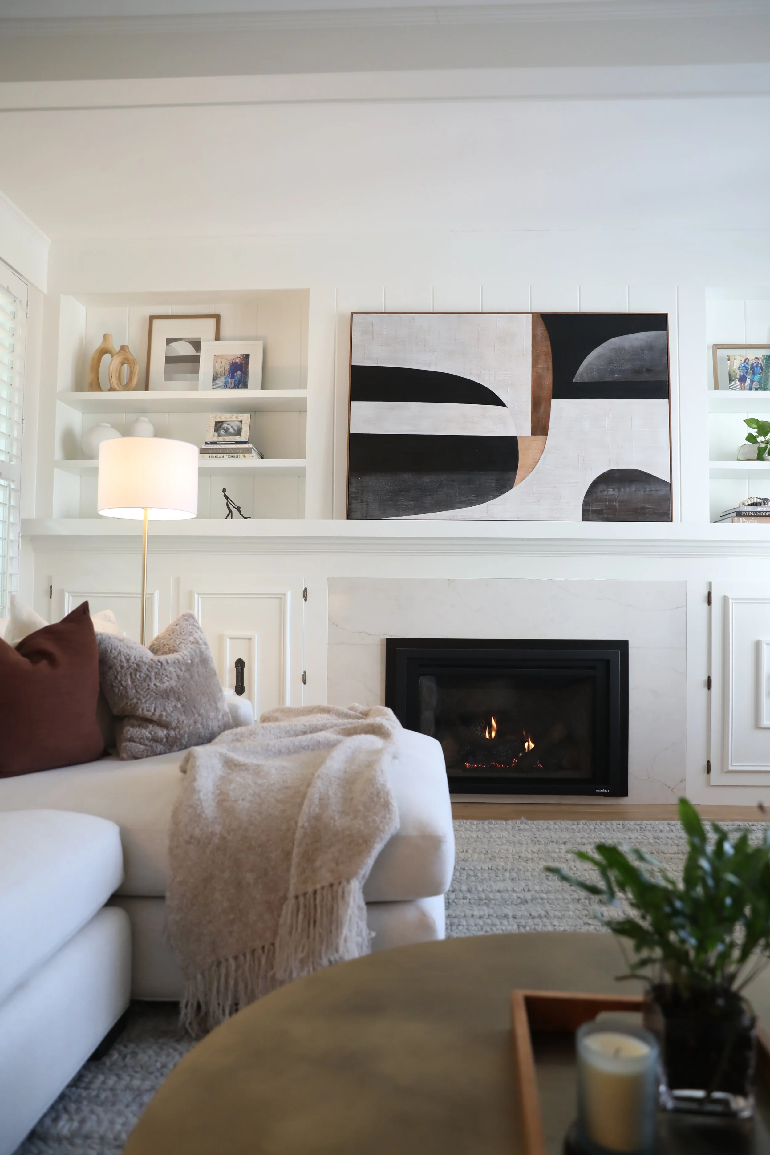 Living room with white sofa, beige throw blanket, and brown pillows. Fireplace with candles and a plant on the mantle. Abstract wall art and framed photographs on white built-in shelves. Lamp illuminating space, and a dark wooden coffee table in the foreground.
