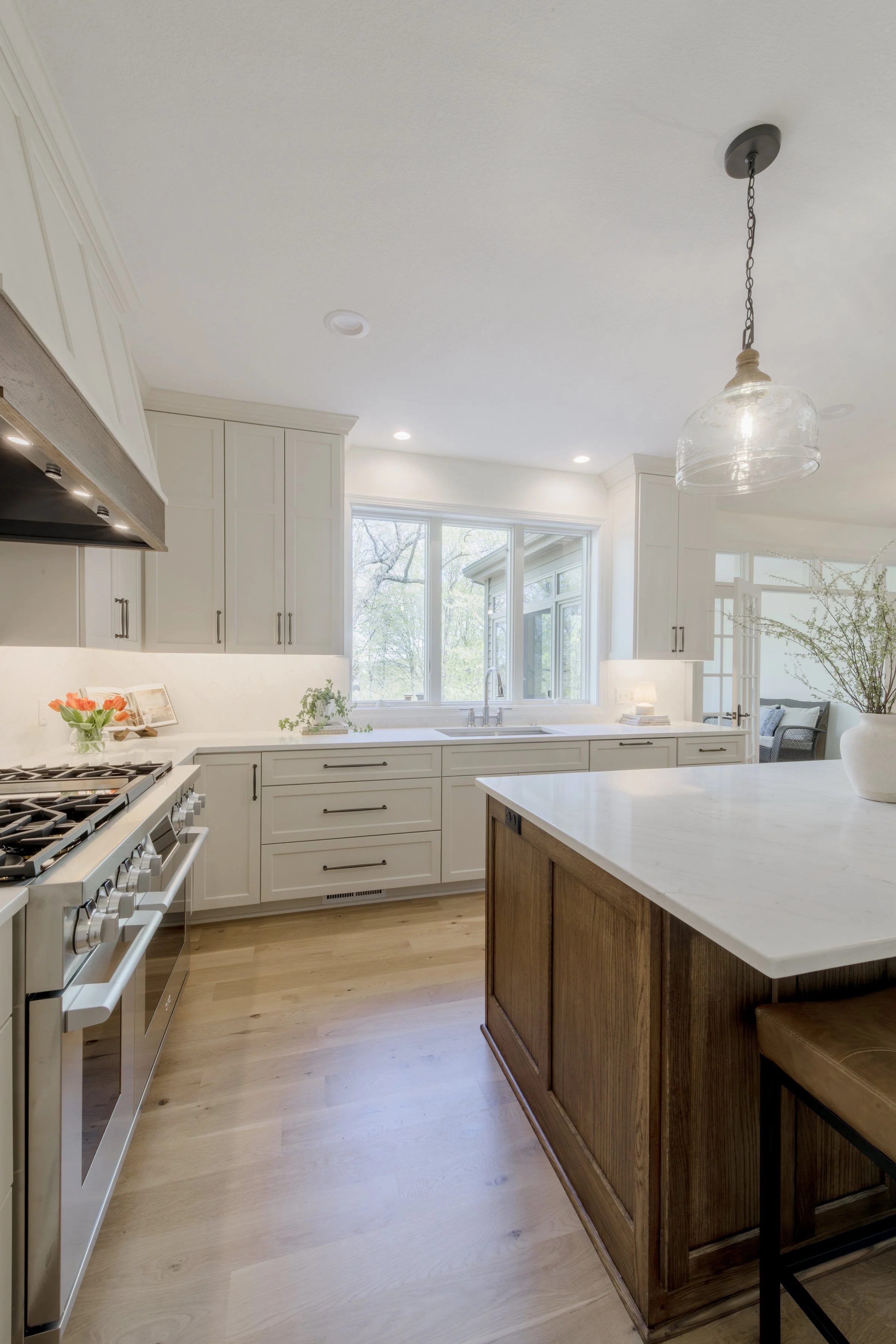 A spacious, bright kitchen with white cabinetry, a stainless steel stove, a large window over the sink, and a modern hanging light fixture. There is a kitchen island with a wooden base and a white countertop, and a small flower arrangement on the cou