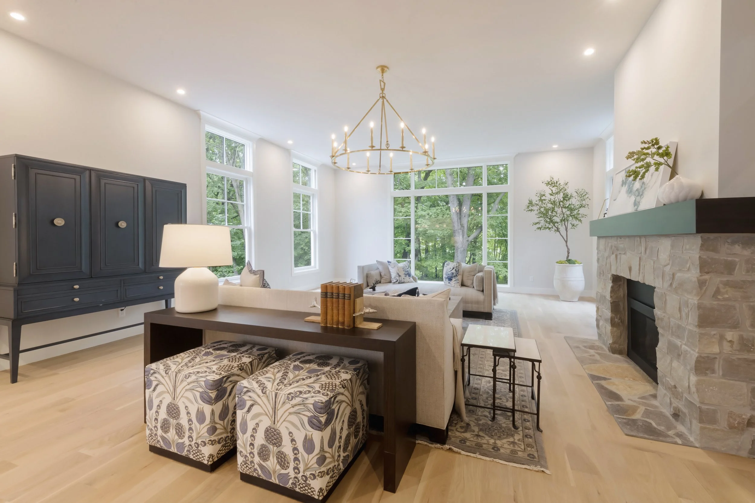 Bright living room with large windows showing green trees outside, featuring a blue cabinet, beige sofas with patterned pillows, a white table lamp, decorative book, a fireplace with a stone mantel, and a chandelier hanging from the ceiling.