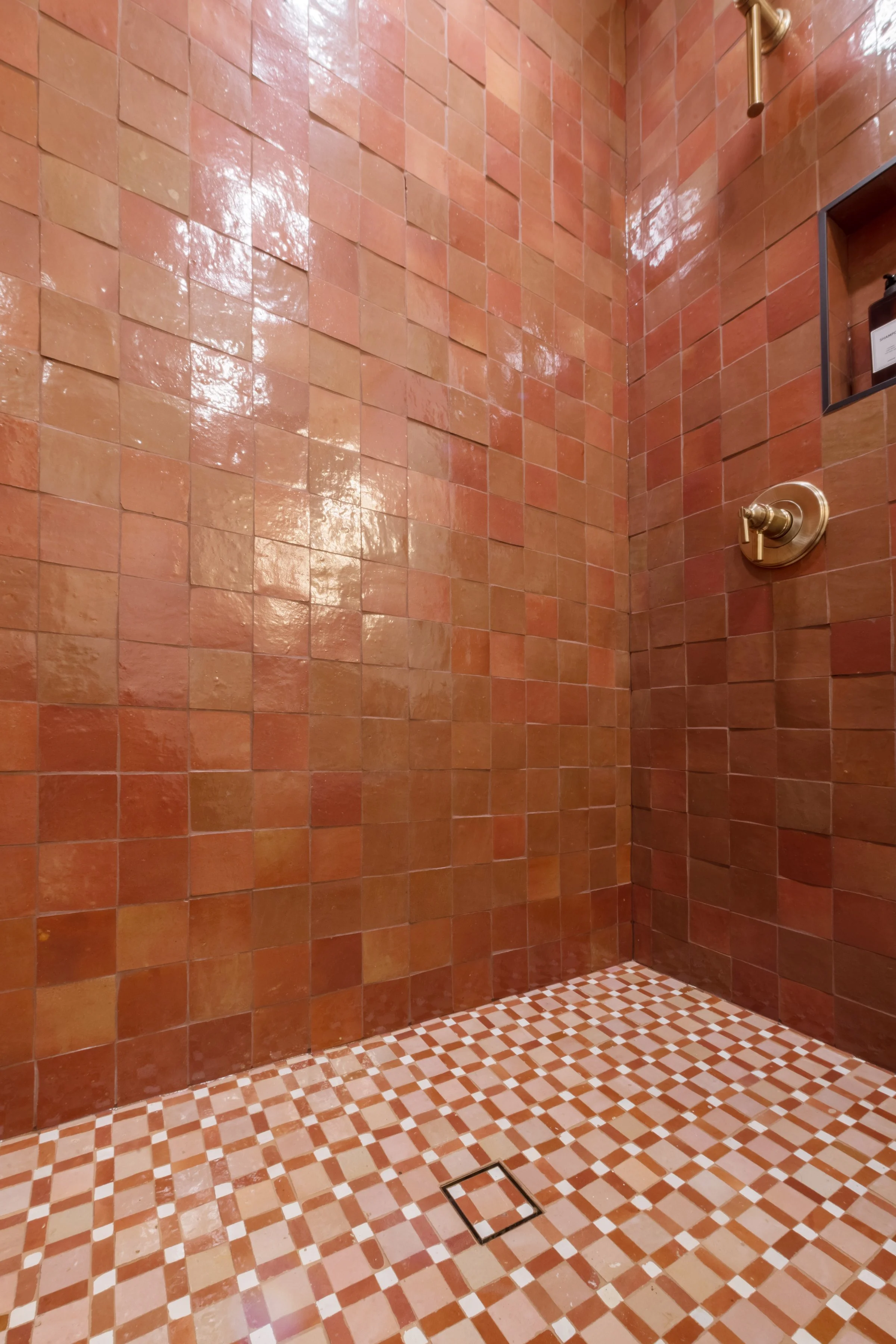 A shower area with orange-brown tiles on the walls and a colorful mosaic tile pattern on the floor, featuring a metal shower head and a control valve.