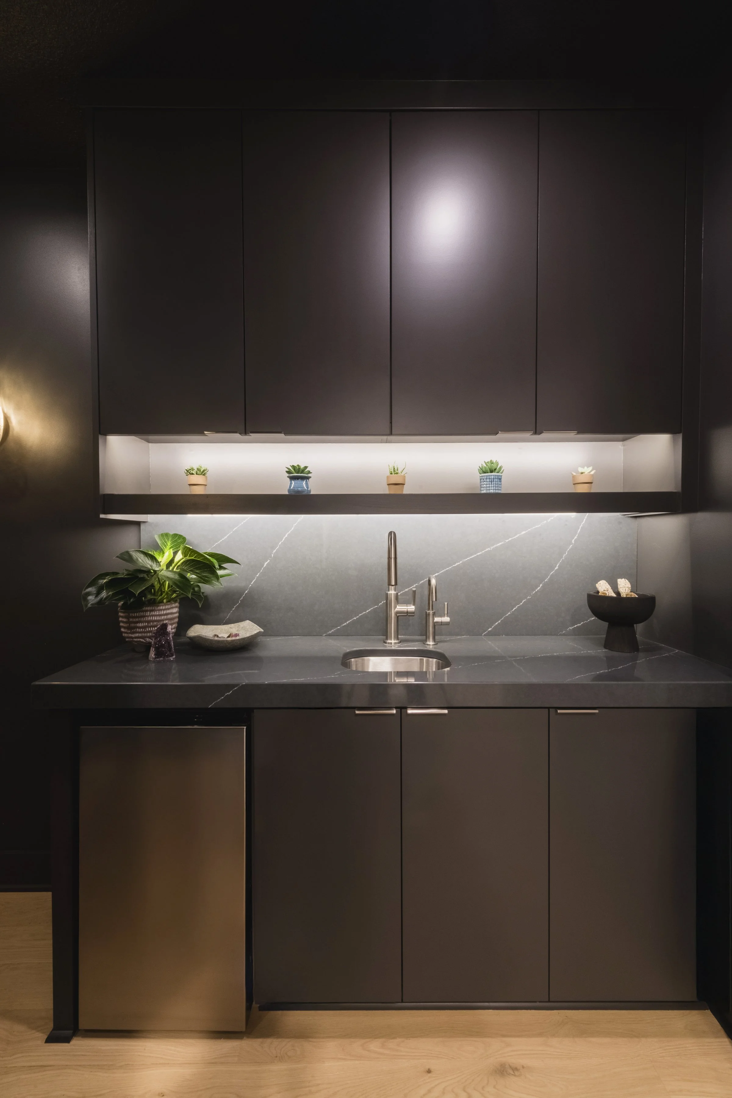 Modern kitchen with black cabinets, gray countertop, stainless steel faucet, green plants, and minimal decor.