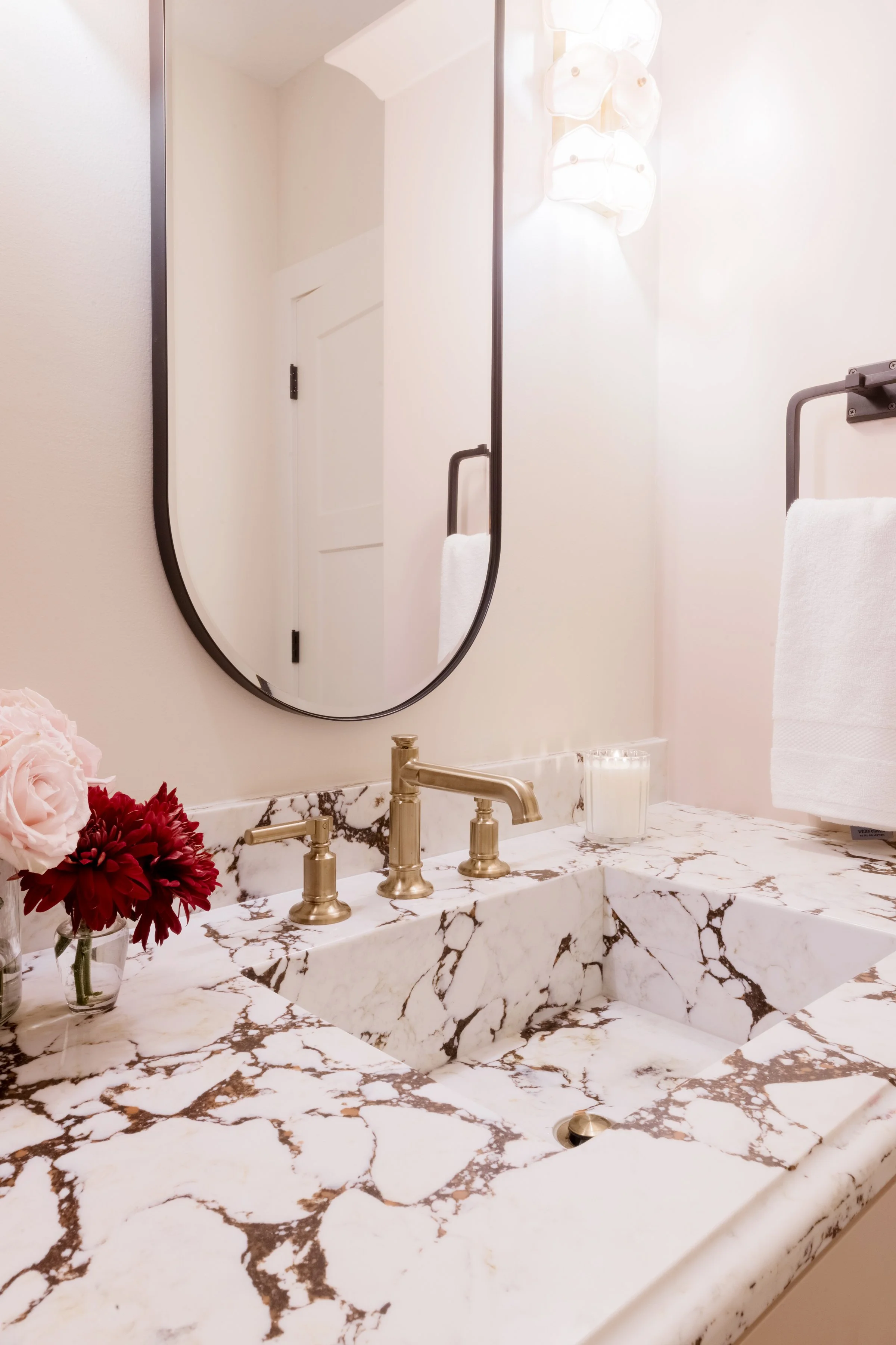 A bathroom vanity with white marble top and a brass faucet, decorated with a bouquet of pink and red flowers, with a wall-mounted mirror, light fixture, and white towel.