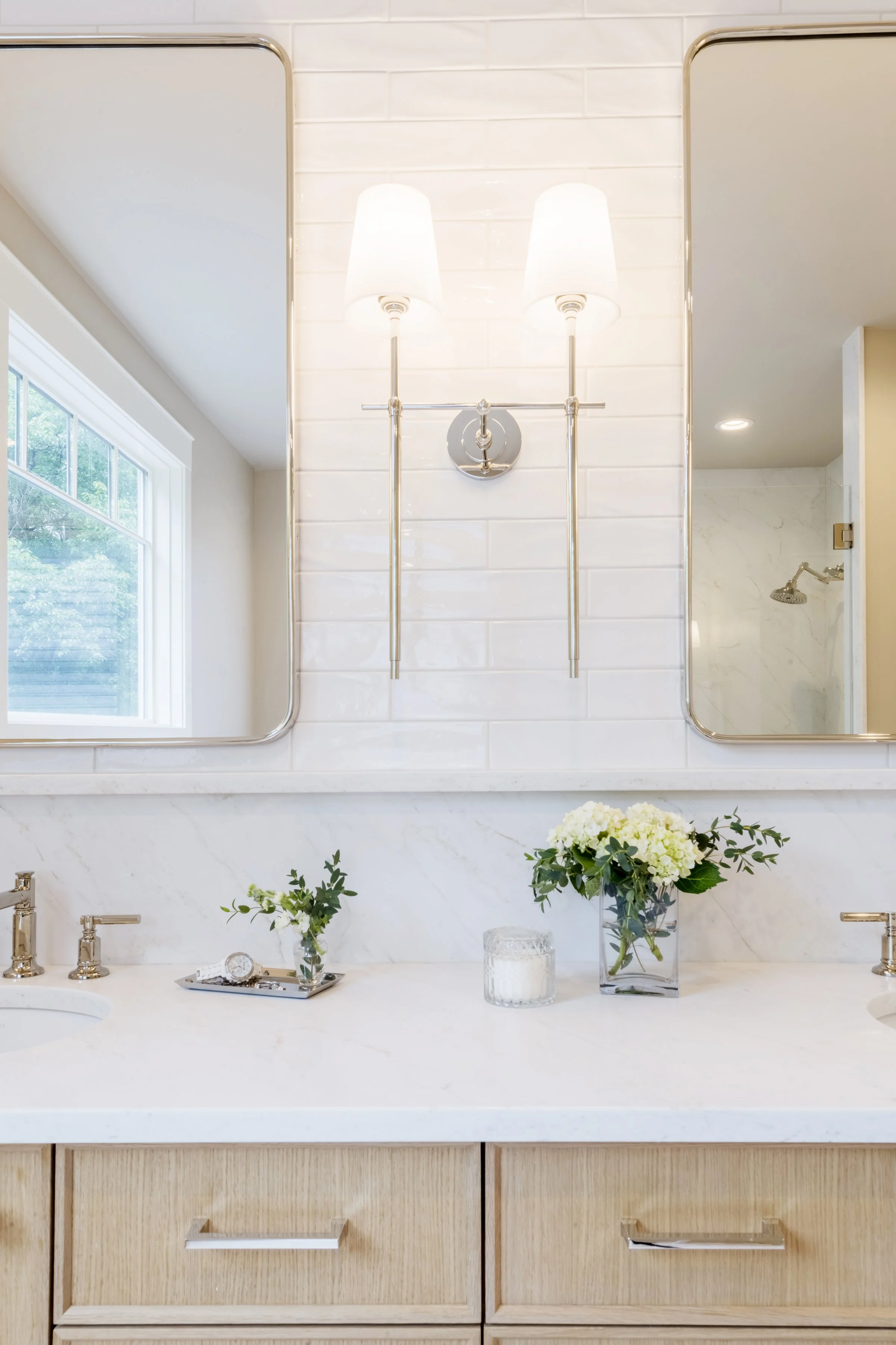 Bathroom vanity with a white marble countertop, two undermount sinks, and layered mirrors. Decor includes two small flower arrangements, a tray with jewelry, and a glass vase with white flowers. A window and shower with a rainfall showerhead are in t