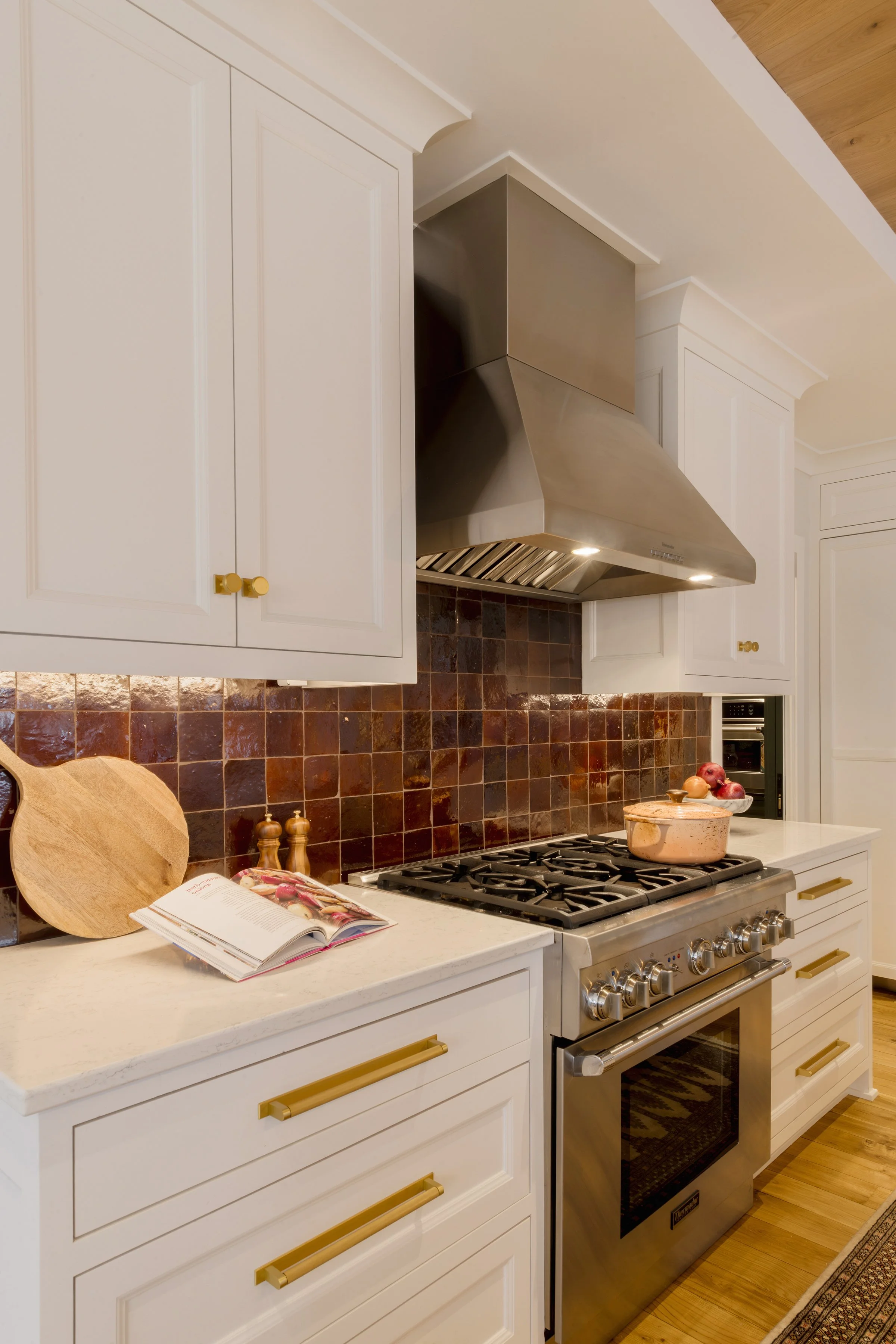 Kitchen with white cabinets with gold handles, a stainless steel range hood over a stove, a cookbook and cutting board on the countertop, and a bowl of apples.