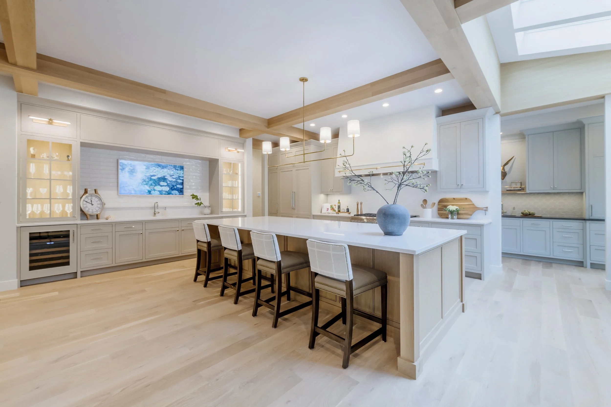Modern kitchen with white cabinets, a large island with four chairs, a vase with branches on the island, and built-in lighting with wooden beams on the ceiling.