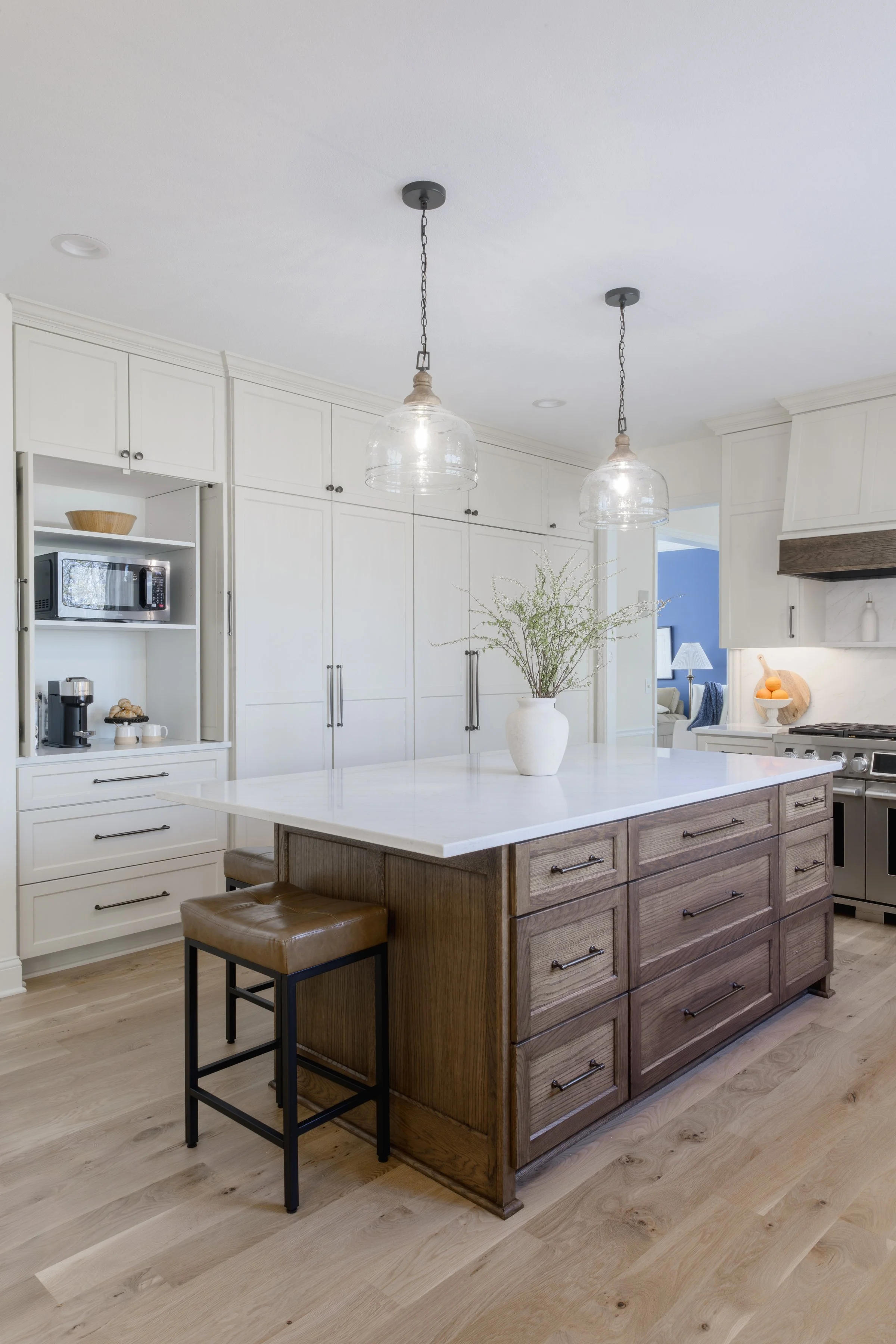 A modern kitchen featuring a white countertop island with dark wood cabinetry, white upper cabinets, pendant lights, and a vase with greenery. The kitchen has light-colored wooden flooring and various appliances and decor.