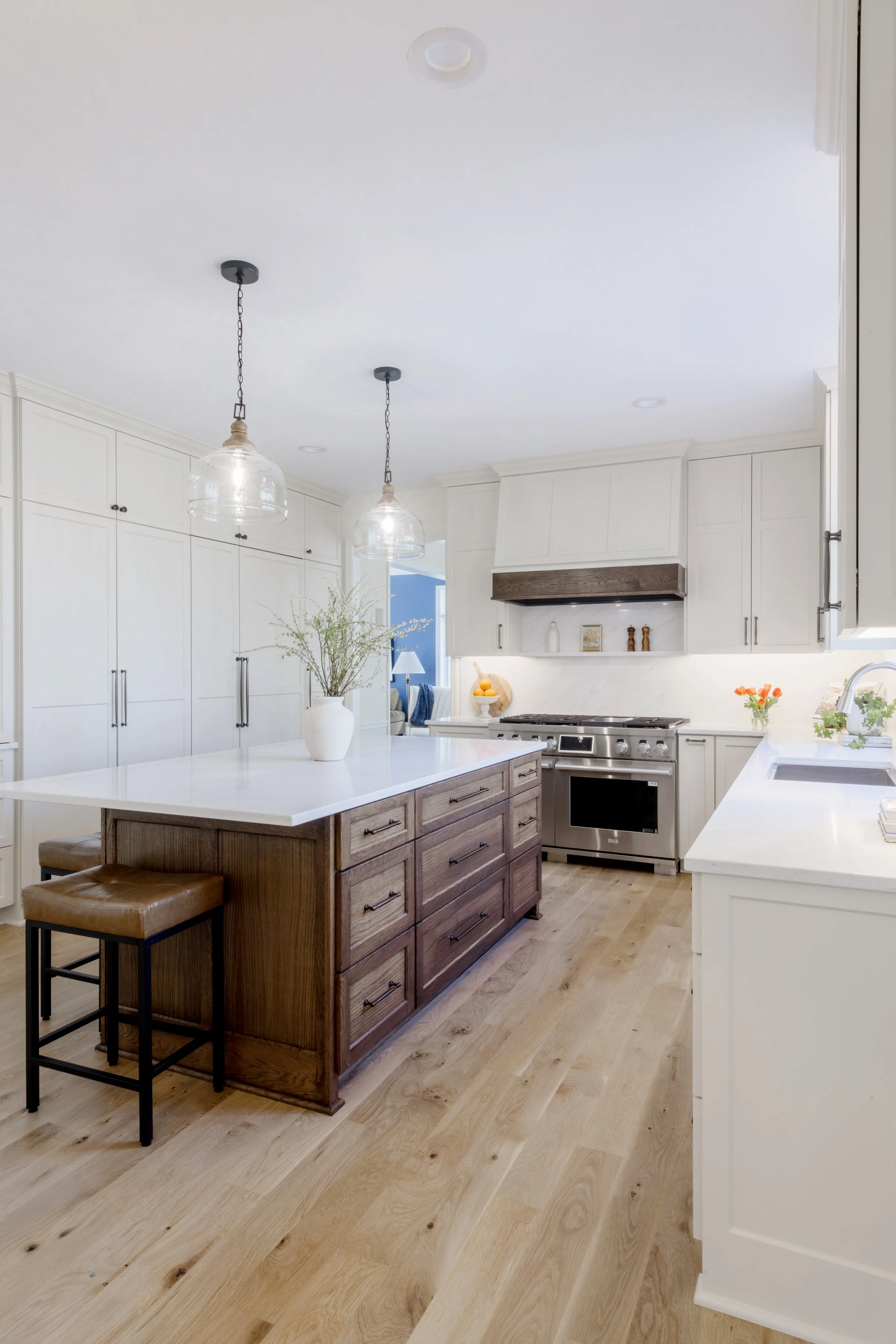 A modern kitchen with white cabinets, a wooden island with a white countertop, and light hardwood flooring. Two pendant lights hang from the ceiling above the island. There are decorative items and a plant on the island, and the kitchen has a stove and floral decorations.