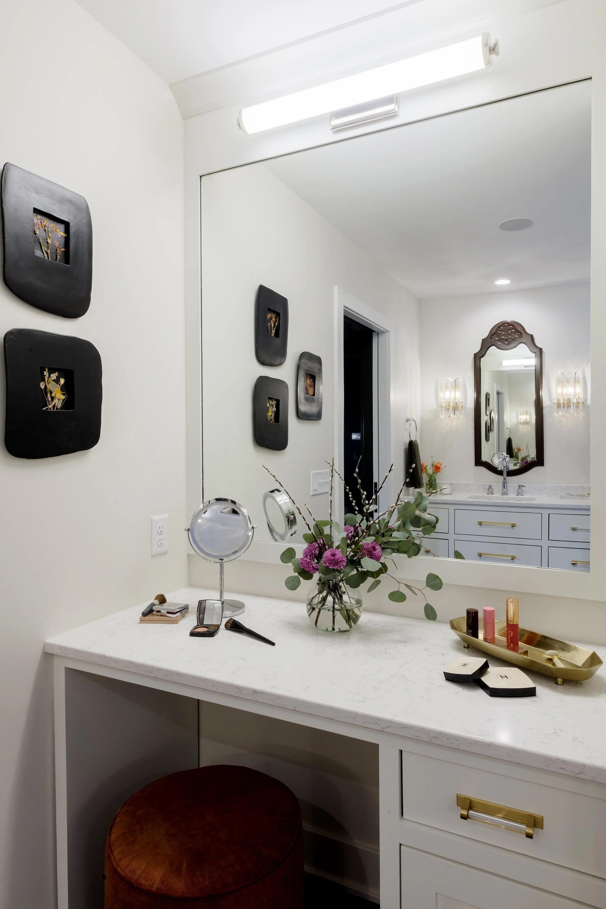A bedroom vanity area with a white marble countertop, a large mirror, and wall-mounted lights. The countertop has makeup items, a round mirrored stand, and a gold tray with lipstick and perfume bottles. A vase with pink flowers and greenery decorates