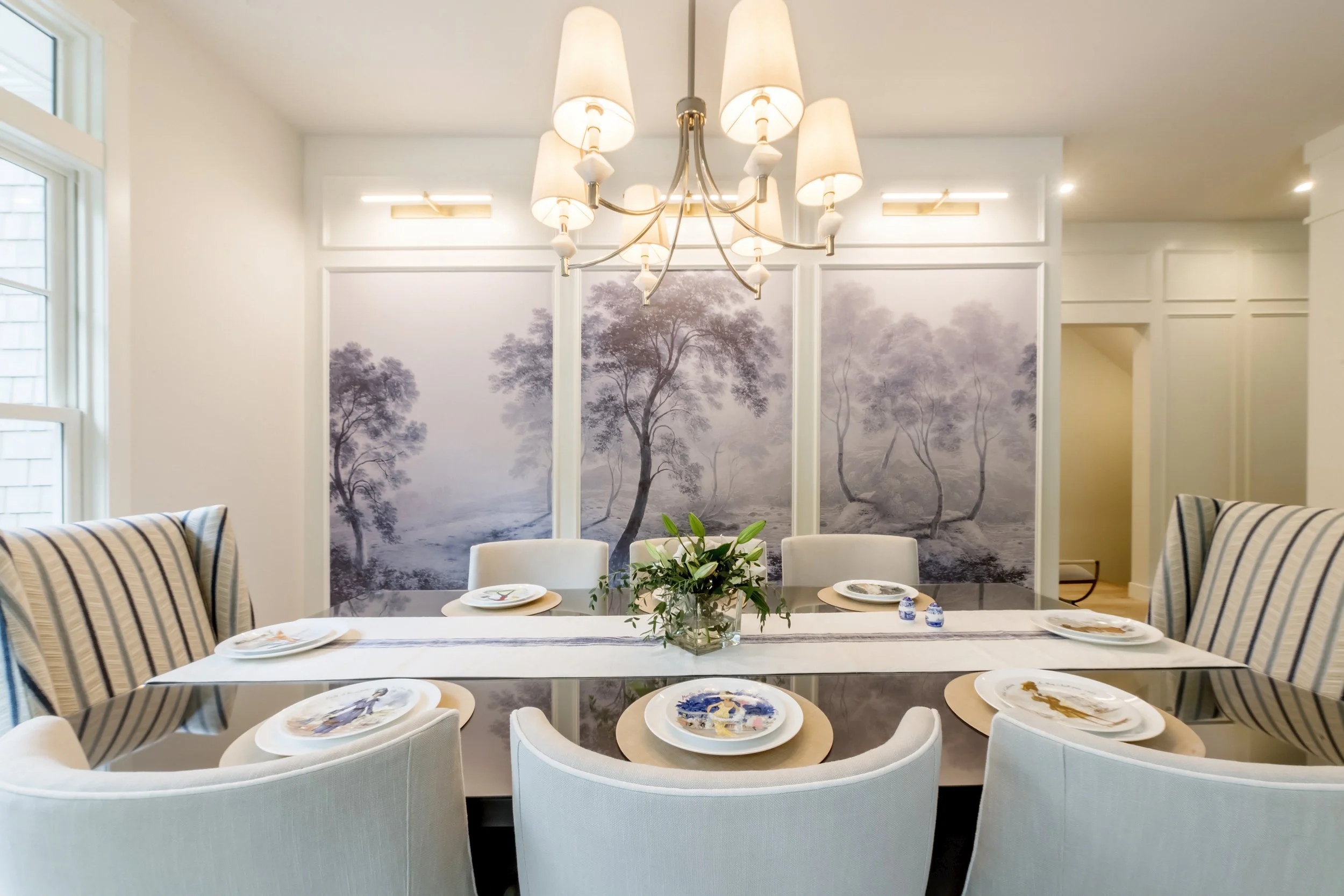 A dining room with a glass table set with plates, a centerpiece, and chairs with striped upholstery. A large wall panel featuring a misty landscape with trees decorates the wall behind the table. A chandelier with multiple lamps hangs from the ceilin