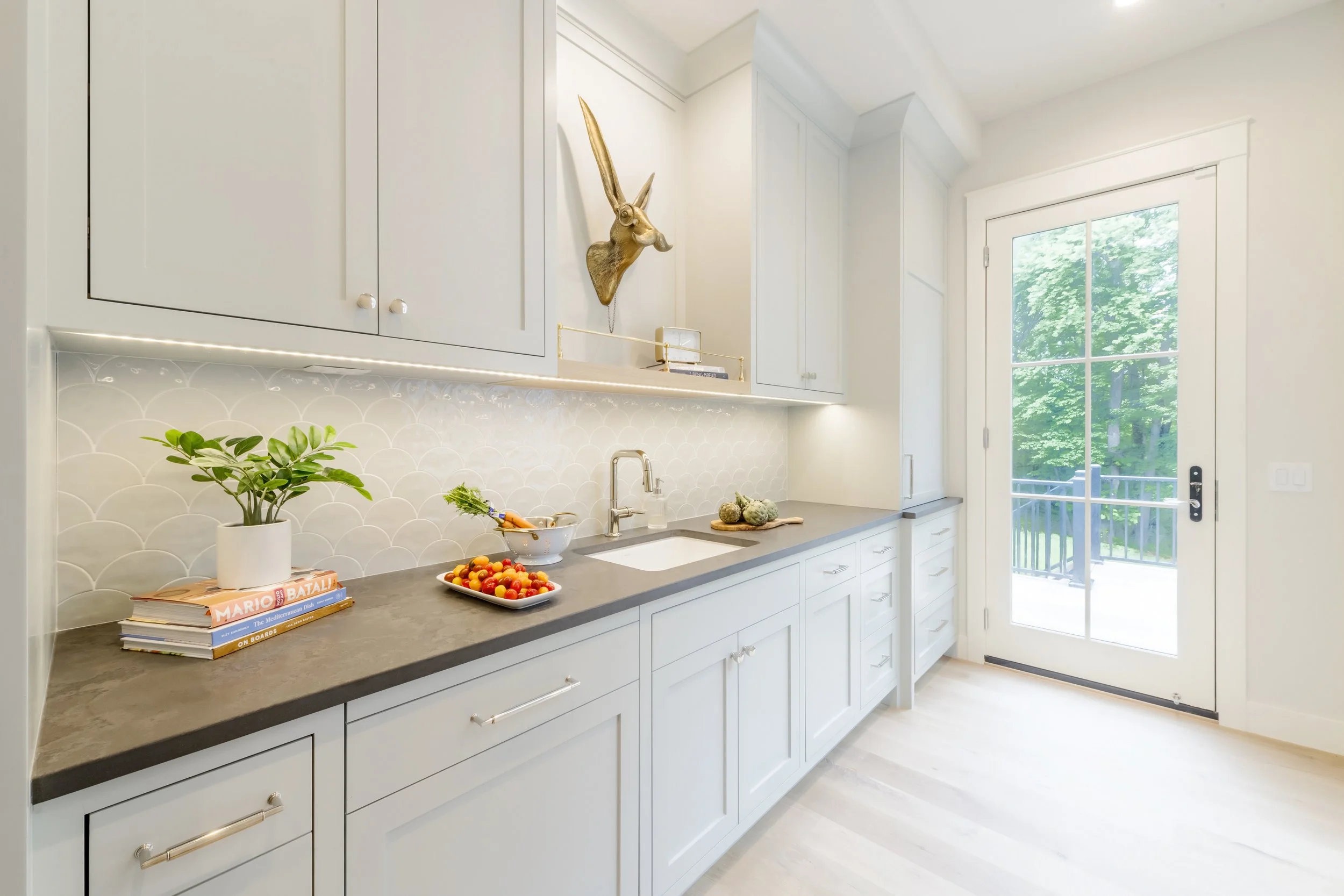 Modern white kitchen with gray countertop, potted plant, books, bowl of cherry tomatoes, and window door showing green trees outside.