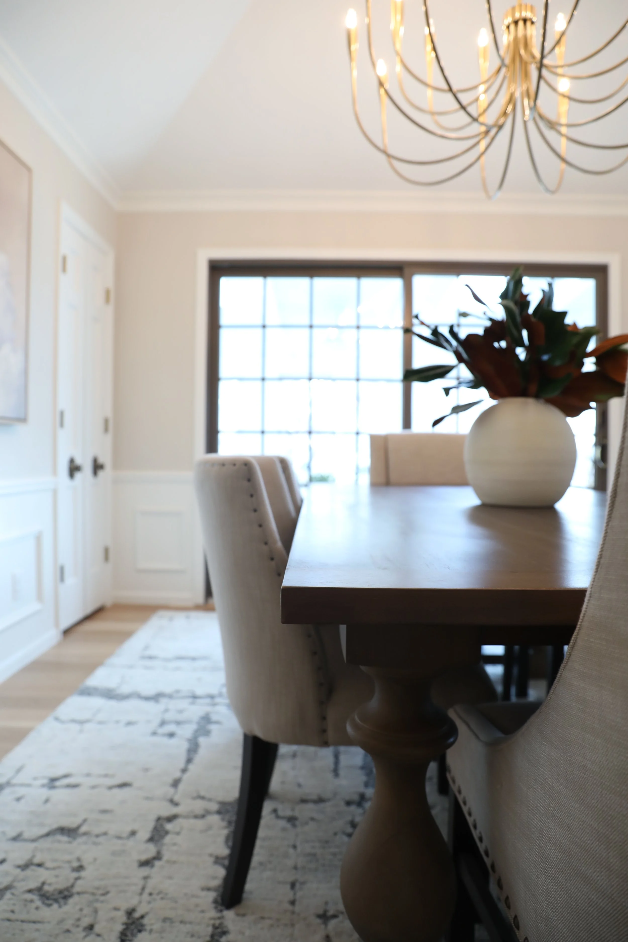 Bright dining room with a wooden table, beige upholstered chairs, decorative beige vase with green and orange flowers, large window with grid pattern, and modern chandelier with exposed bulbs.