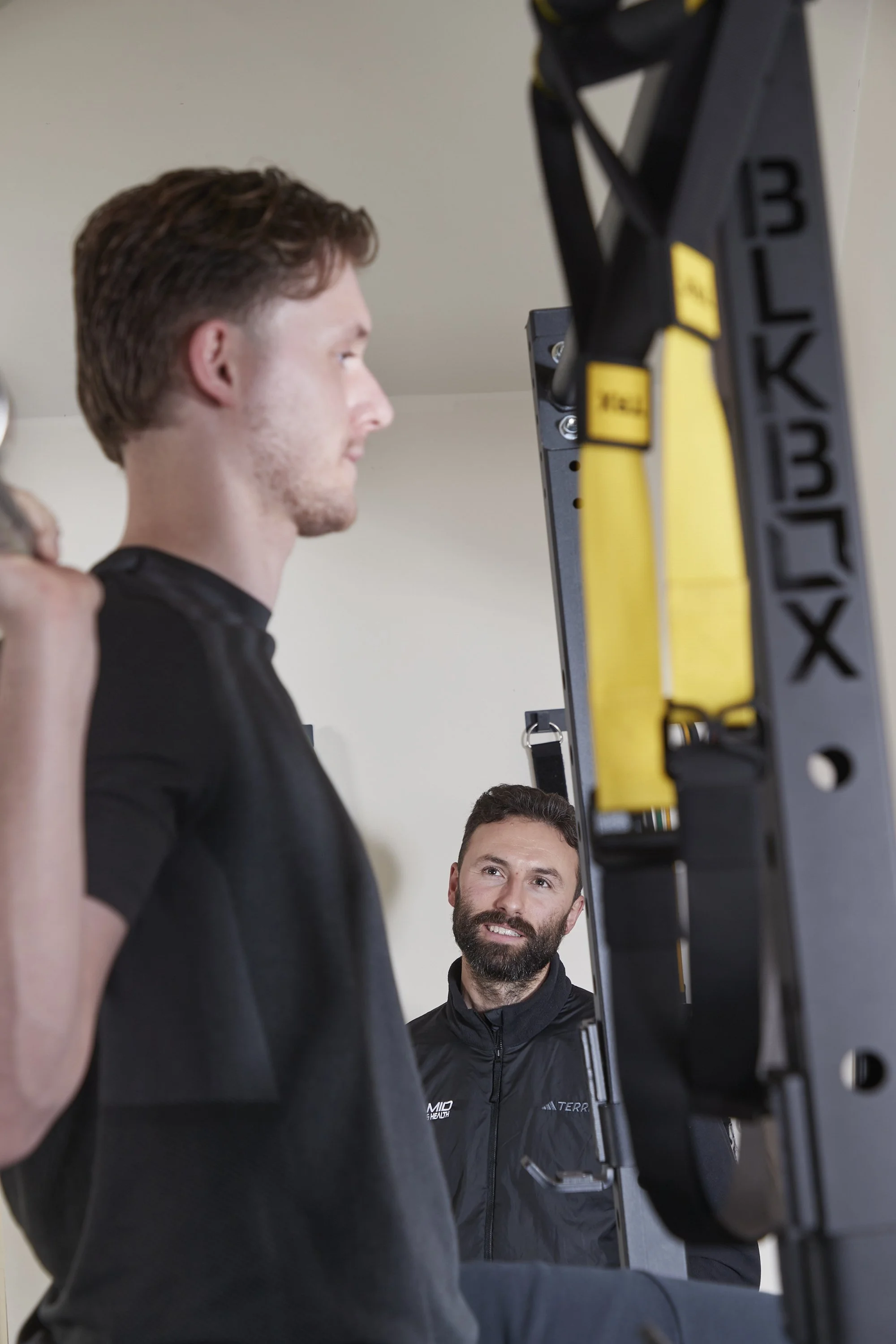 Two men in athletic wear in a gym, one with a beard and one clean-shaven, looking at a piece of fitness equipment or machinery.