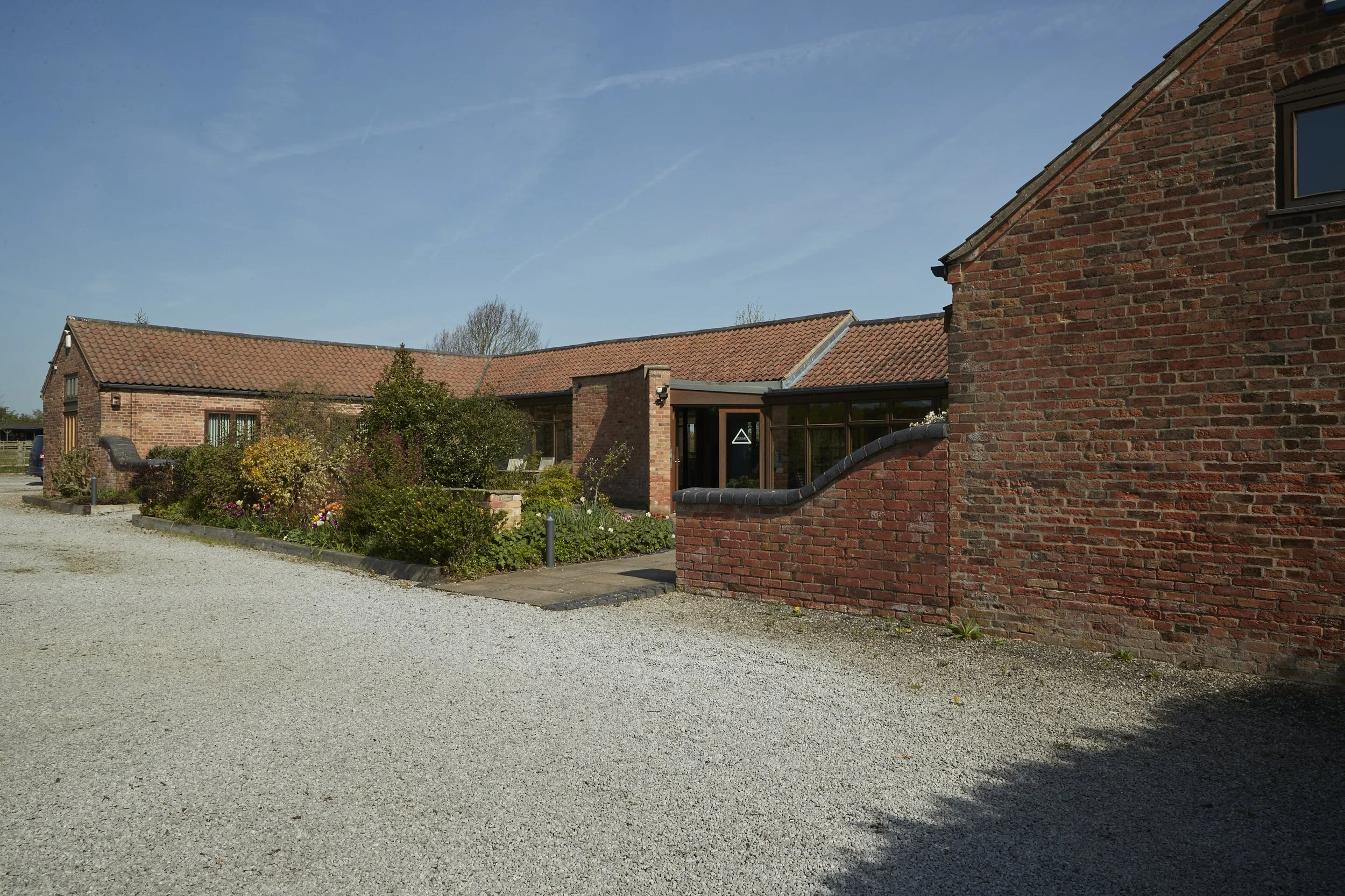 A brick building with a red tile roof and a garden area in front, under a clear blue sky.