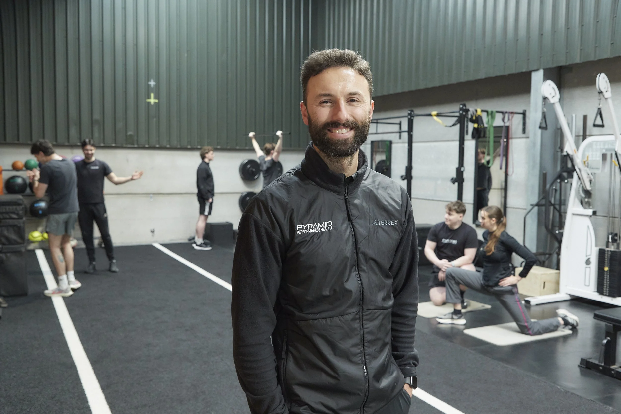 Smiling man with a beard in a black jacket standing in a gym, with people exercising and fitness equipment in the background.