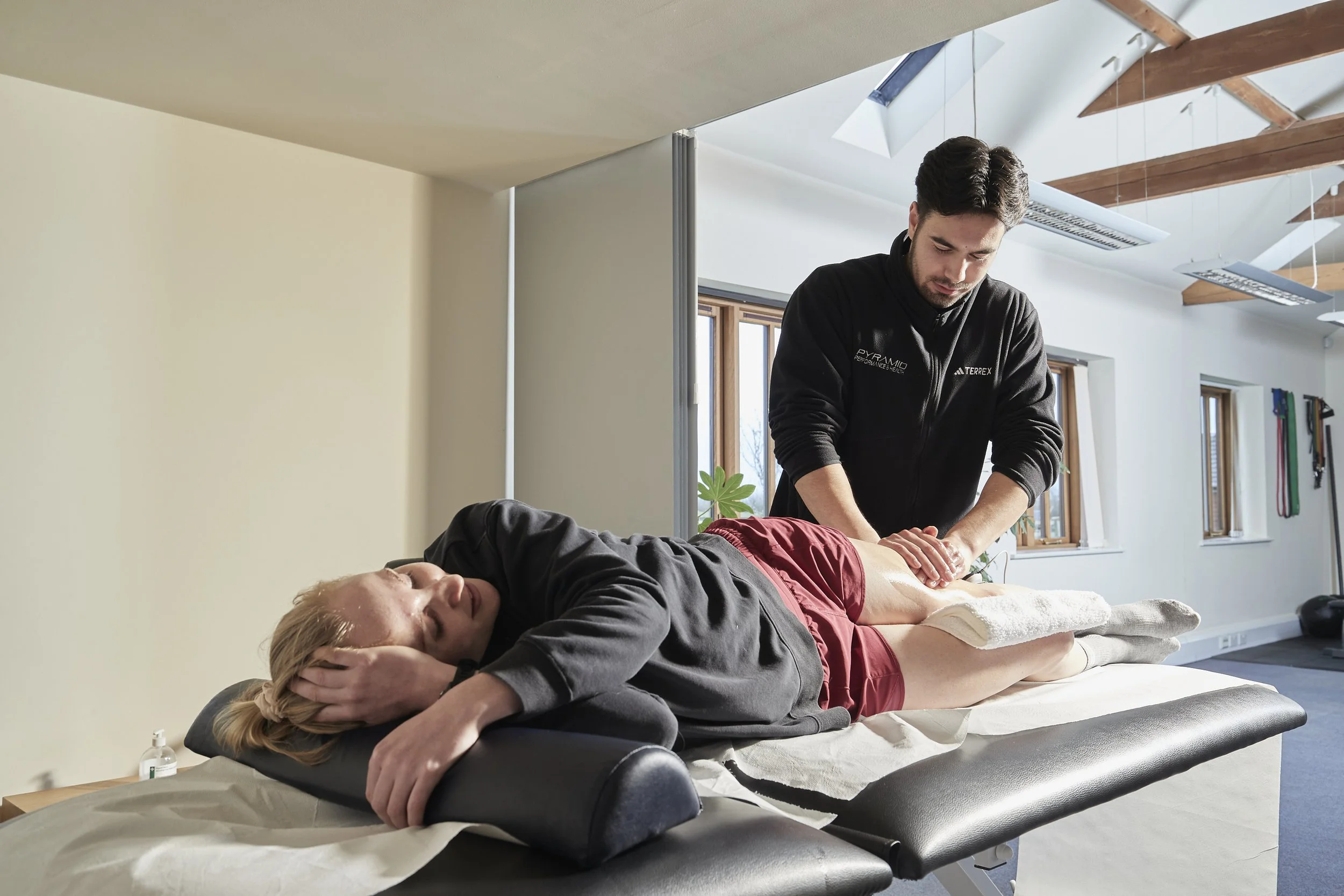 A woman with blonde hair lying face down on a massage table receiving a massage from a man standing beside her in a bright room with large windows and wooden beams. The woman is resting with her hands under her forehead, while the man is focused on m