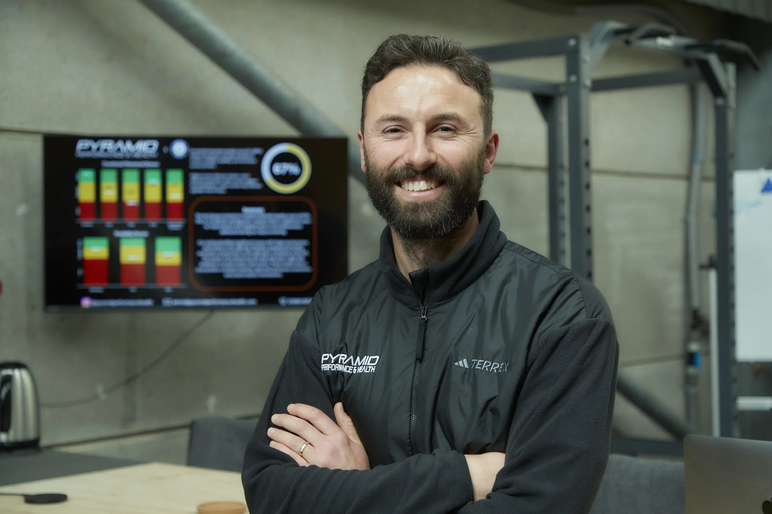 A smiling man with a beard and short dark hair, wearing a black jacket with the words 'PYRAMID PERFORMANCE & HEALTH' and 'TERREX' on it, poses in a gym or fitness center with his arms crossed. Behind him, there is a digital display screen with colorful graphs and information, and exercise equipment can be seen in the background.