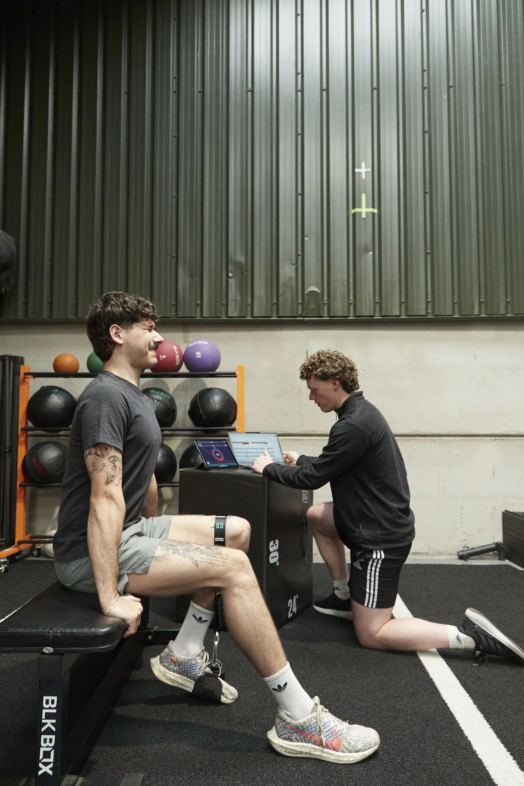 Two men in a gym, with one seated and the other kneeling in front of a black plyometric box. The seated man is smiling and looking up, wearing a black t-shirt, gray shorts, and Nike sneakers. The kneeling man is focused on a device, wearing a black j