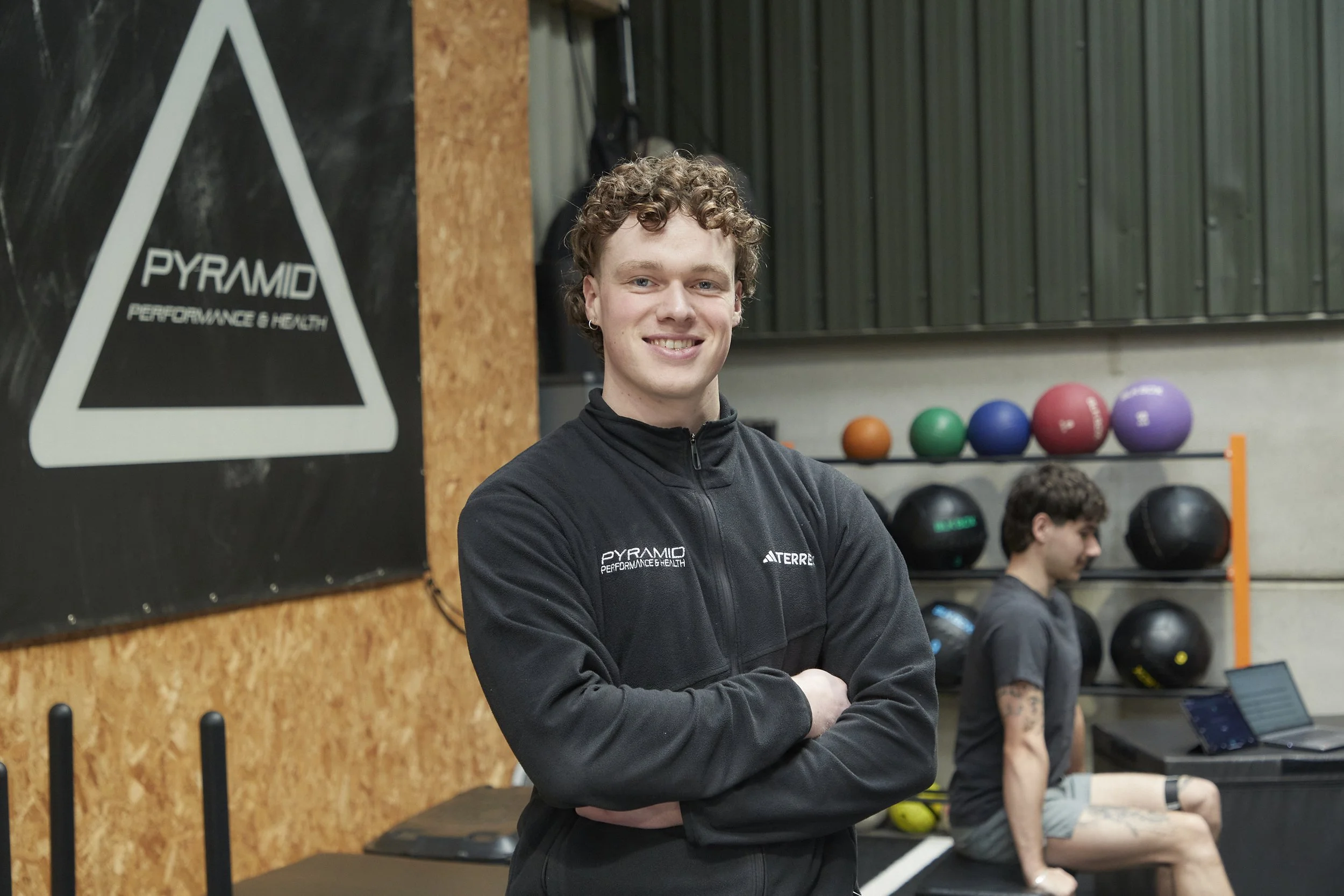 A young man with curly hair and earrings smiling at the camera in a gym setting, surrounded by exercise equipment and weights.
