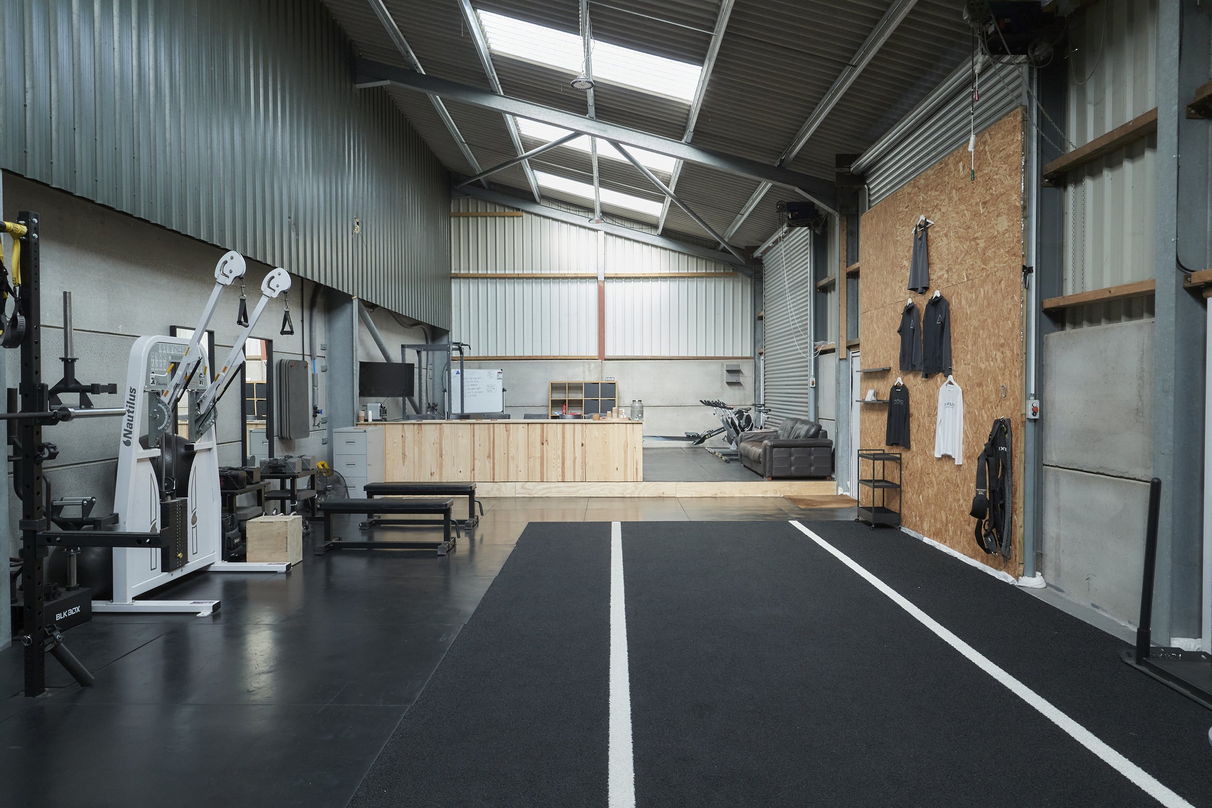 Interior of a gym with workout equipment, a reception desk, and a sitting area, metal walls, and a high ceiling with skylights.