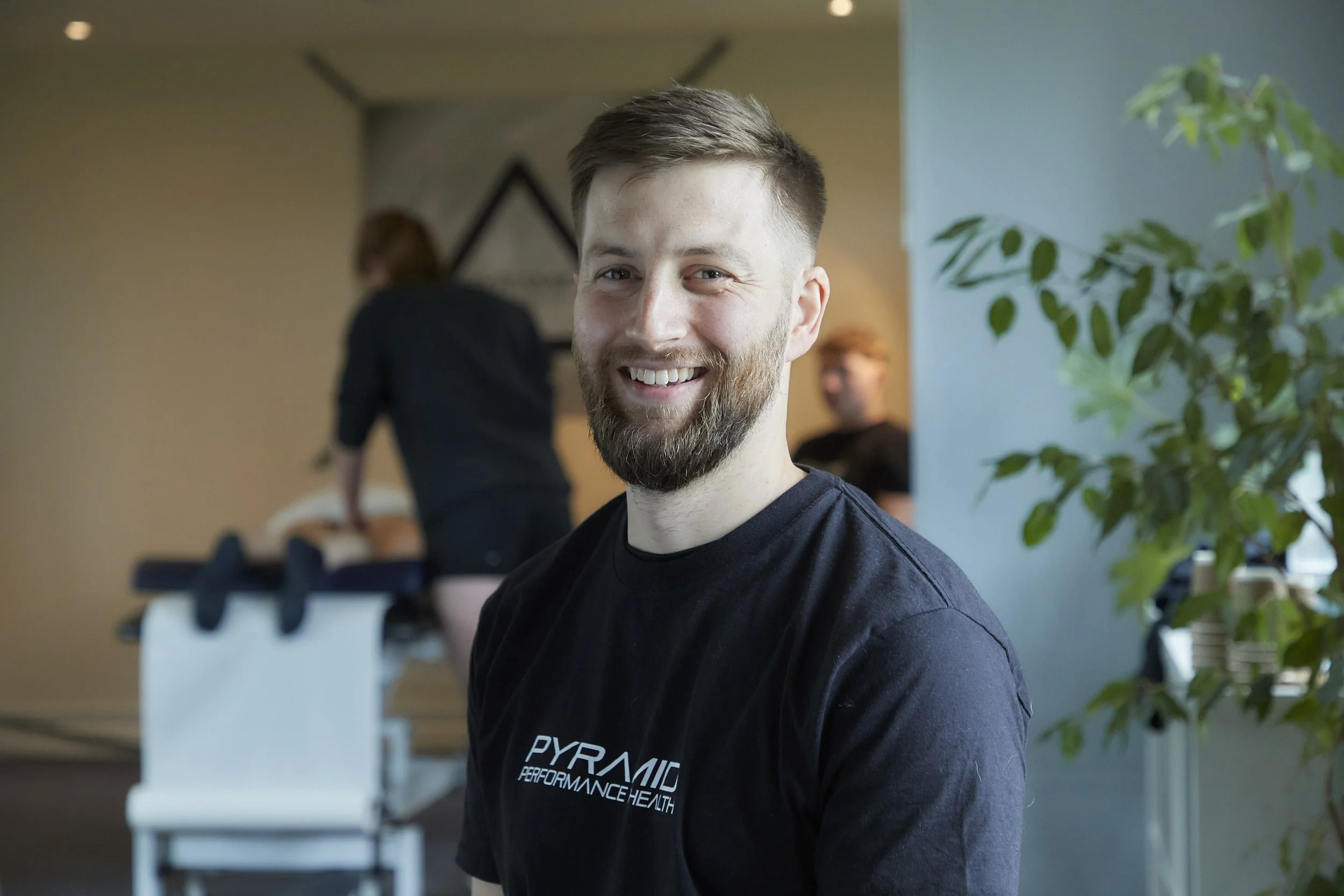 A smiling man with a beard and short hair wearing a black shirt that says 'Pyramid Performance Health', seated indoors near a potted plant. Behind him, a person is working at a counter in a room with soft lighting.