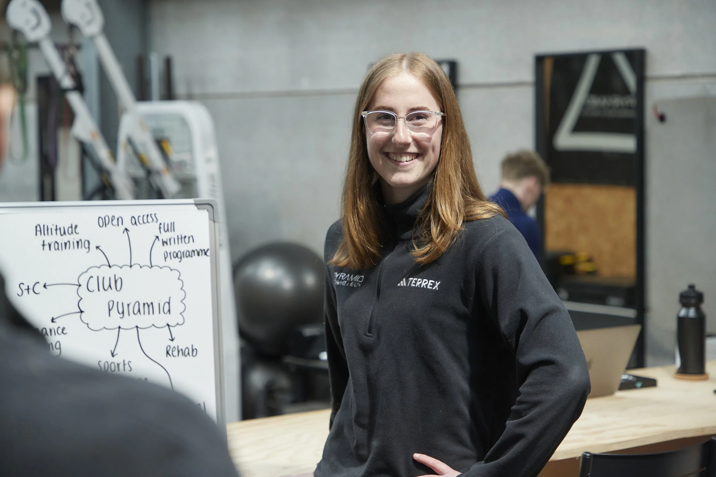 A young woman with red hair, wearing glasses and a black jacket, smiling as she stands in a gym or workout space. In the background, there are gym equipment and a whiteboard with a diagram about a fitness or training program.
