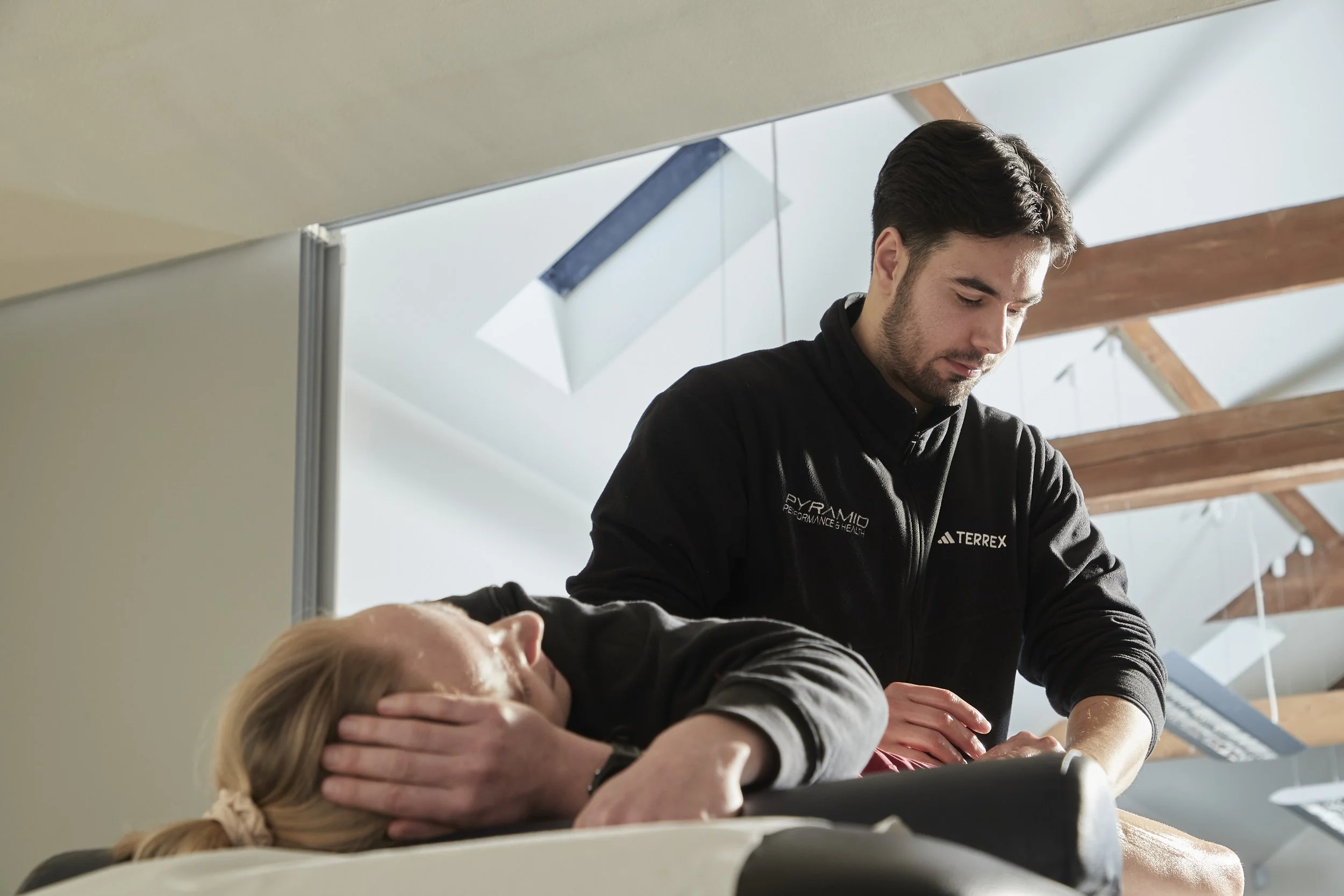 A man with dark hair and beard helping a woman with blonde hair during a physical therapy or medical treatment session. The woman is lying on a treatment table, and the man is pressing on her forehead and neck area. The setting appears to be a modern, well-lit clinic or rehabilitation center with a ceiling featuring wooden beams and skylights.
