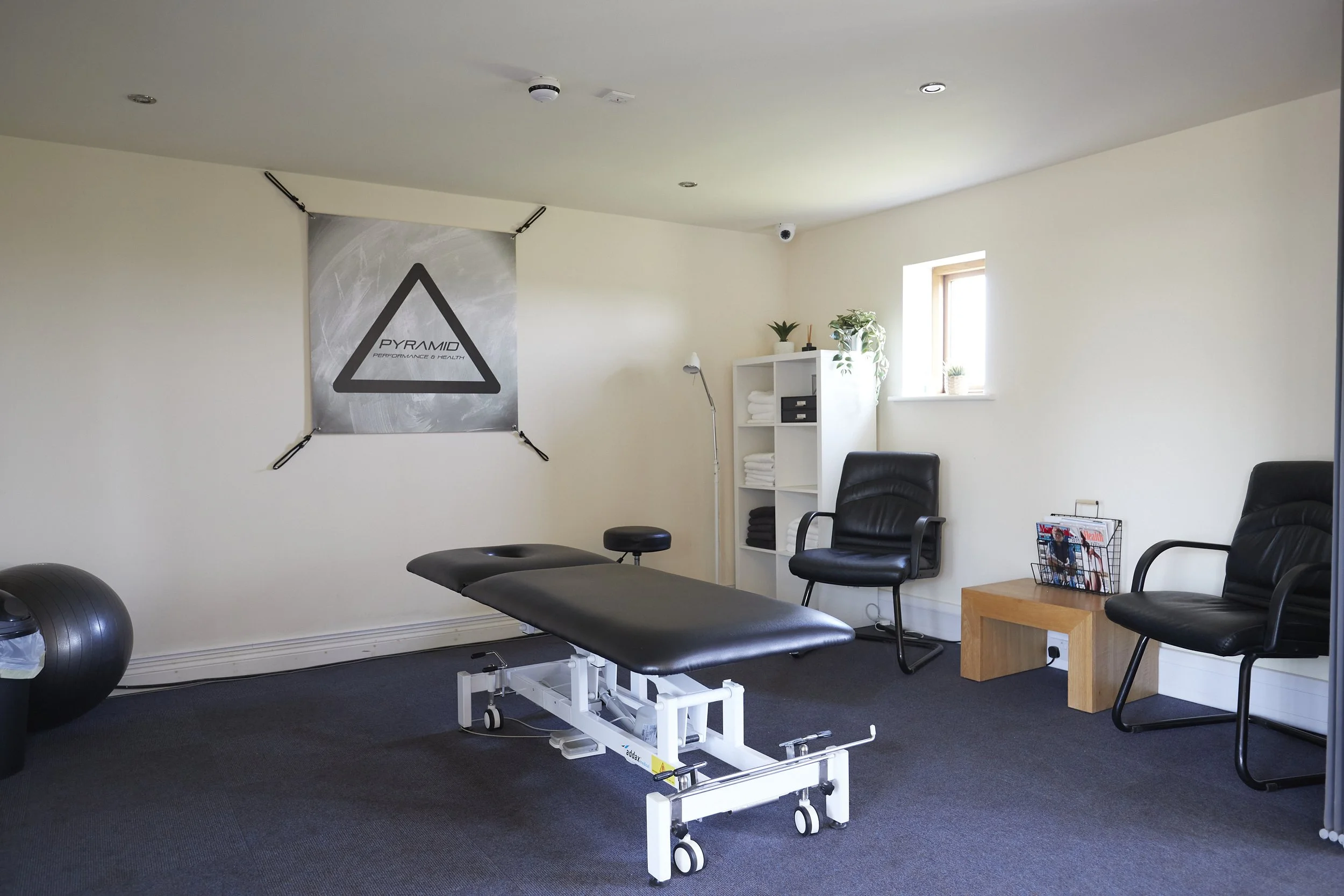 Interior of a fitness or physical therapy room with a massage or therapy table, exercise ball, chairs, bookshelf with towels, potted plants, and a large square window letting in natural light.