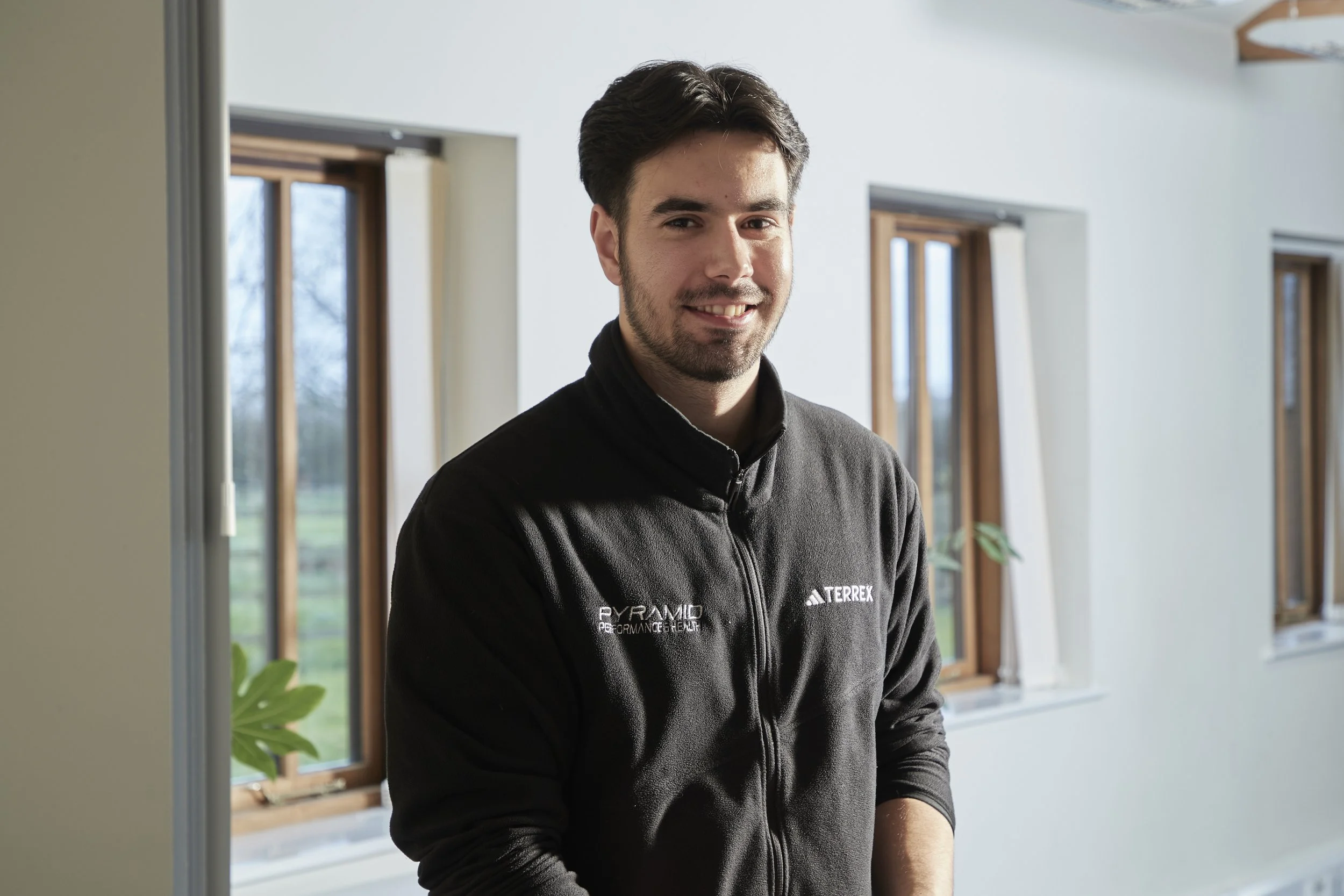 Smiling young man with dark hair and a beard, wearing a black jacket with logos, standing indoors near windows, with a plant partially visible on the left.
