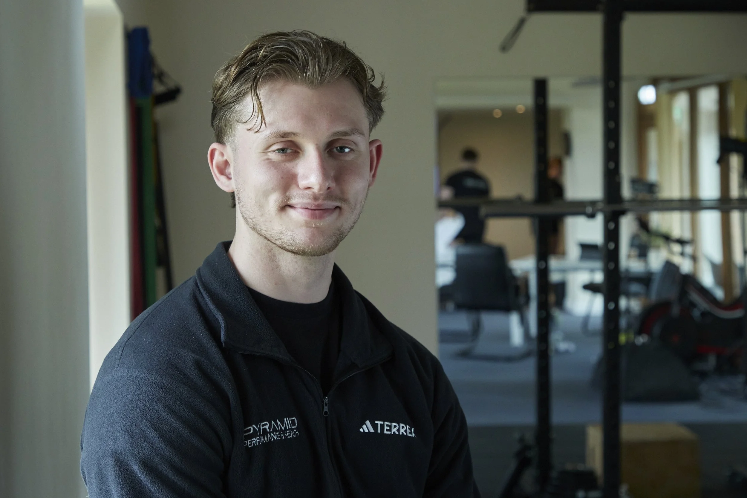 A young man with light brown hair and blue eyes smiling at the camera, wearing a black jacket with white logos, standing in a gym or fitness center.