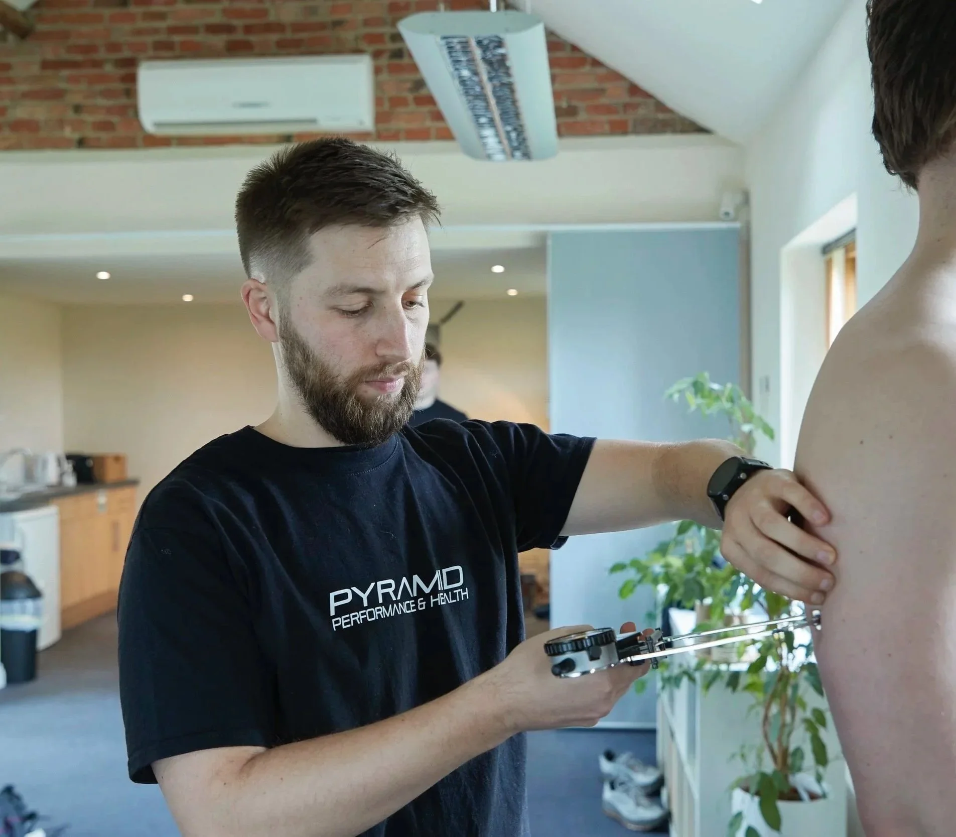 Man in black t-shirt with 'PYRAMID PERFORMANCE & HEALTH' on it, working on a person's shoulder using equipment, in a well-lit room with brick wall, air conditioner, and plants.