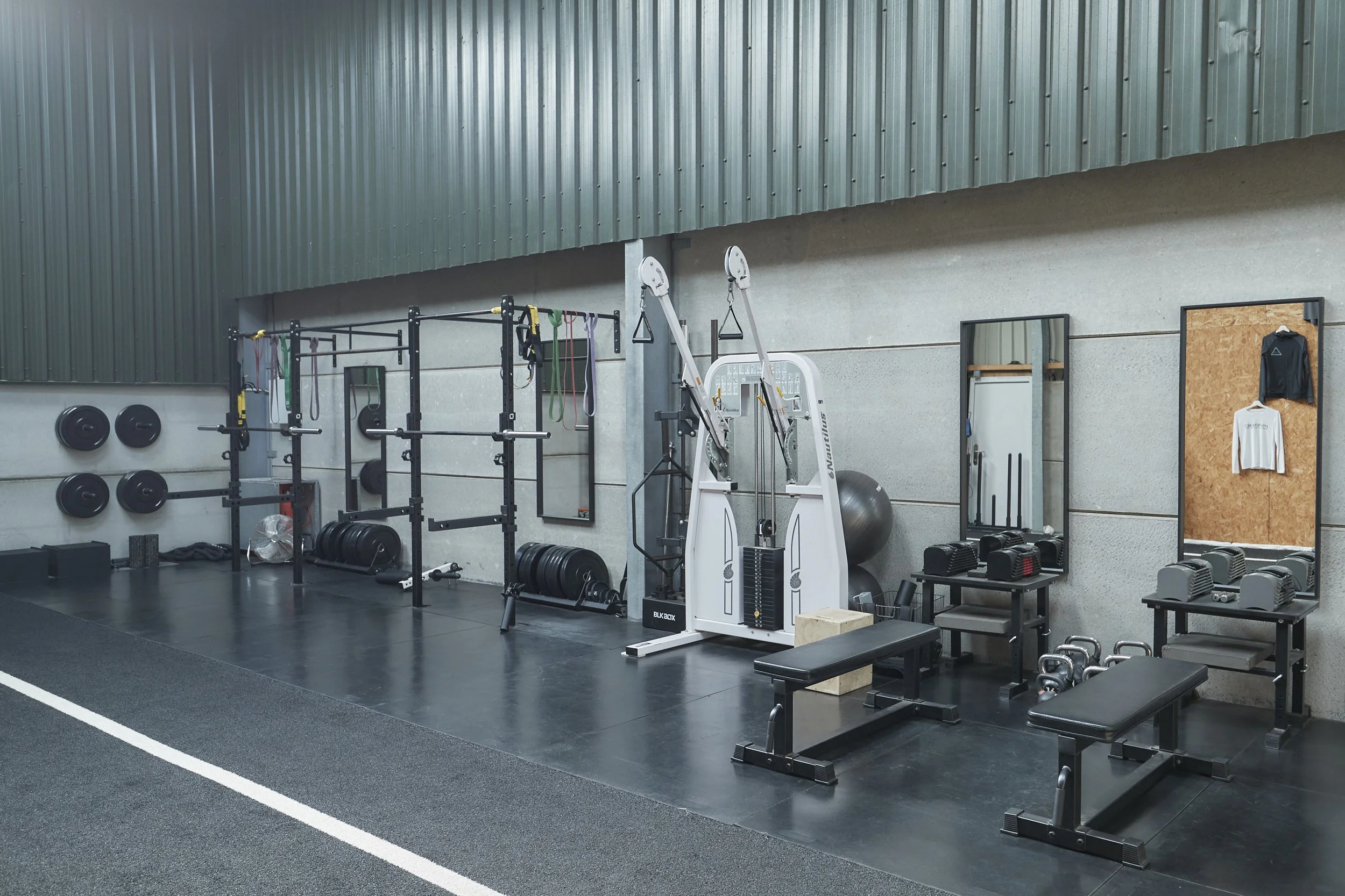 Gym with black rubber flooring, metal wall panels, and workout equipment including weights, resistance bands, a white strength machine, medicine balls, wall mirrors, and clothing hanging on the wall.