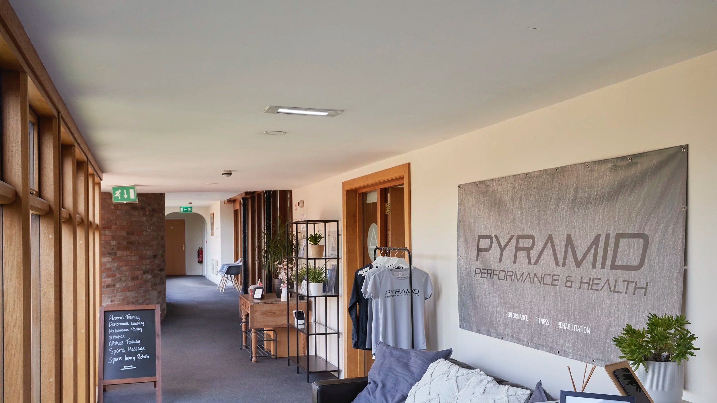 Indoor reception area with wooden accents, brick wall, promotional clothing on a rack, plants, and performance and health banners.