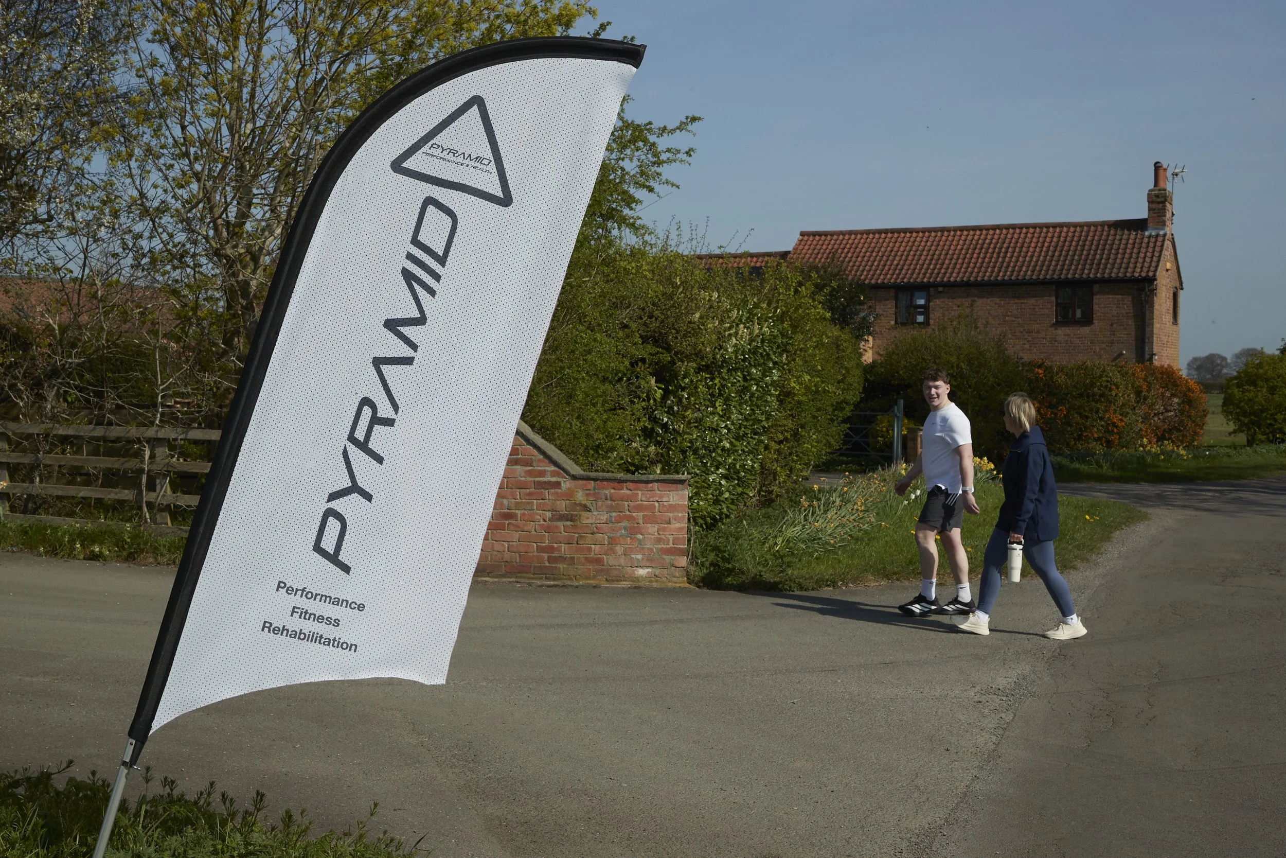 A white feather-shaped banner with black text and logo stands on a sidewalk near a grassy area. Two people are walking past it, one with a white shirt and shorts, and the other with a blue jacket and dark pants. In the background, there is a red bric