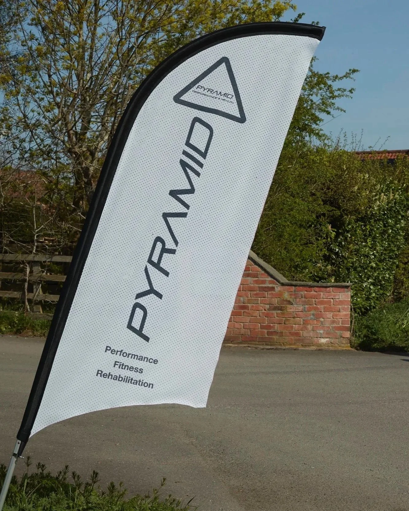 A white feather banner with black border, displaying the logo 'PYRAMID' and words 'Performance', 'Fitness', 'Rehabilitation' on a street with a brick wall and trees in the background.